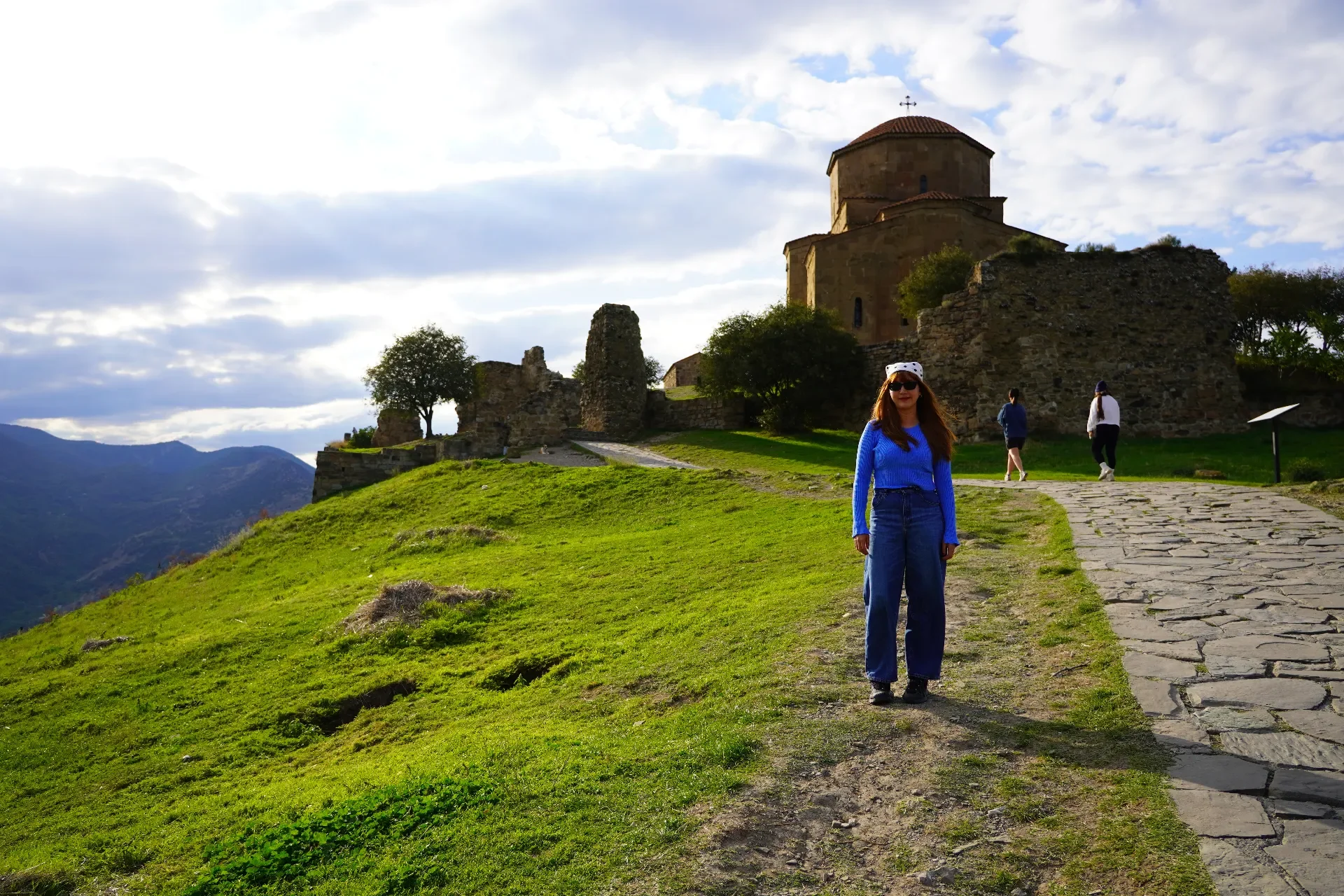 A woman in a blue sweater and jeans walking on a grassy path in front of an ancient stone church and ruins, with a cloudy sky and mountain landscape in the background.