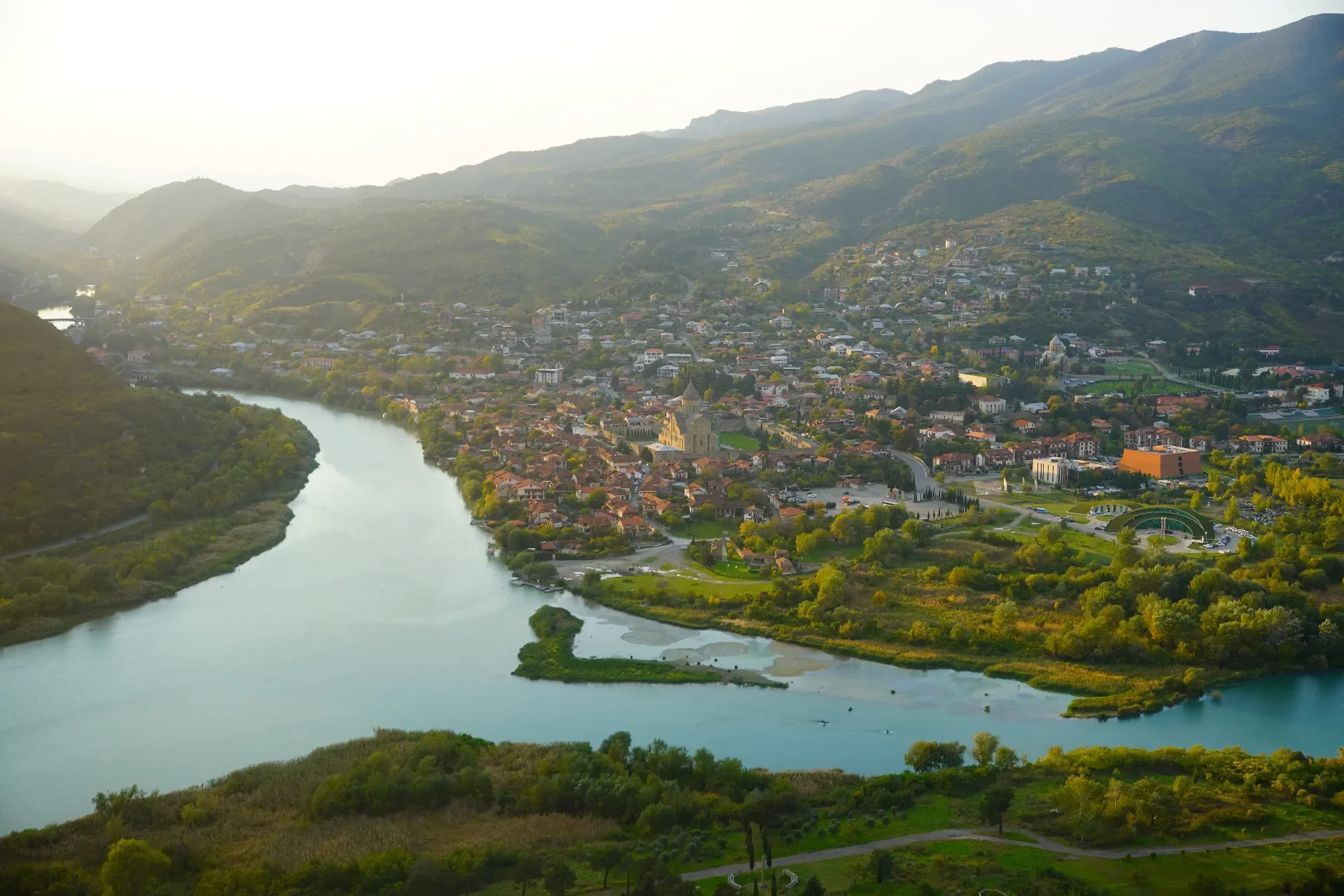 Aerial view of a town surrounded by greenery and mountains, with a winding river flowing through it, during daylight hours.