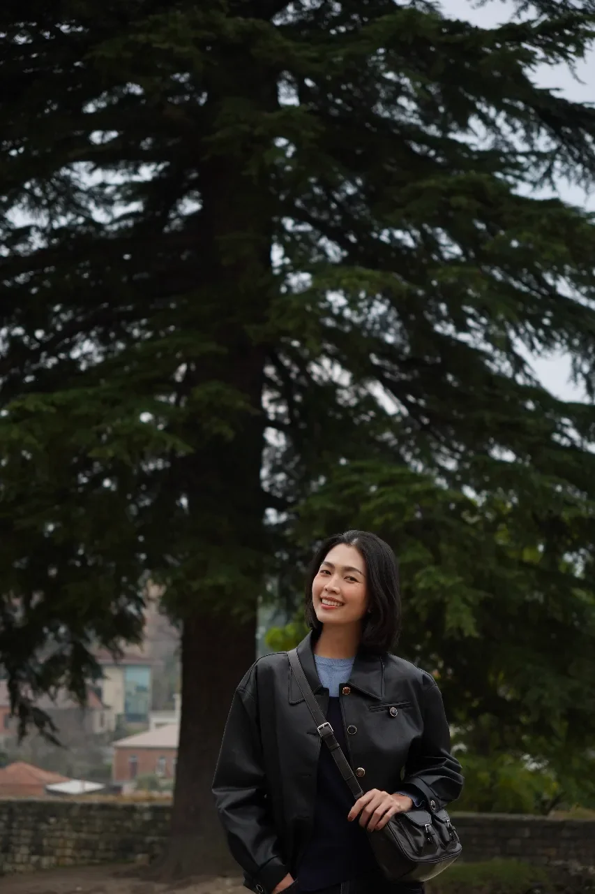 A young woman with shoulder-length black hair smiling and standing outdoors in front of a large evergreen tree, wearing a black jacket and carrying a black purse.