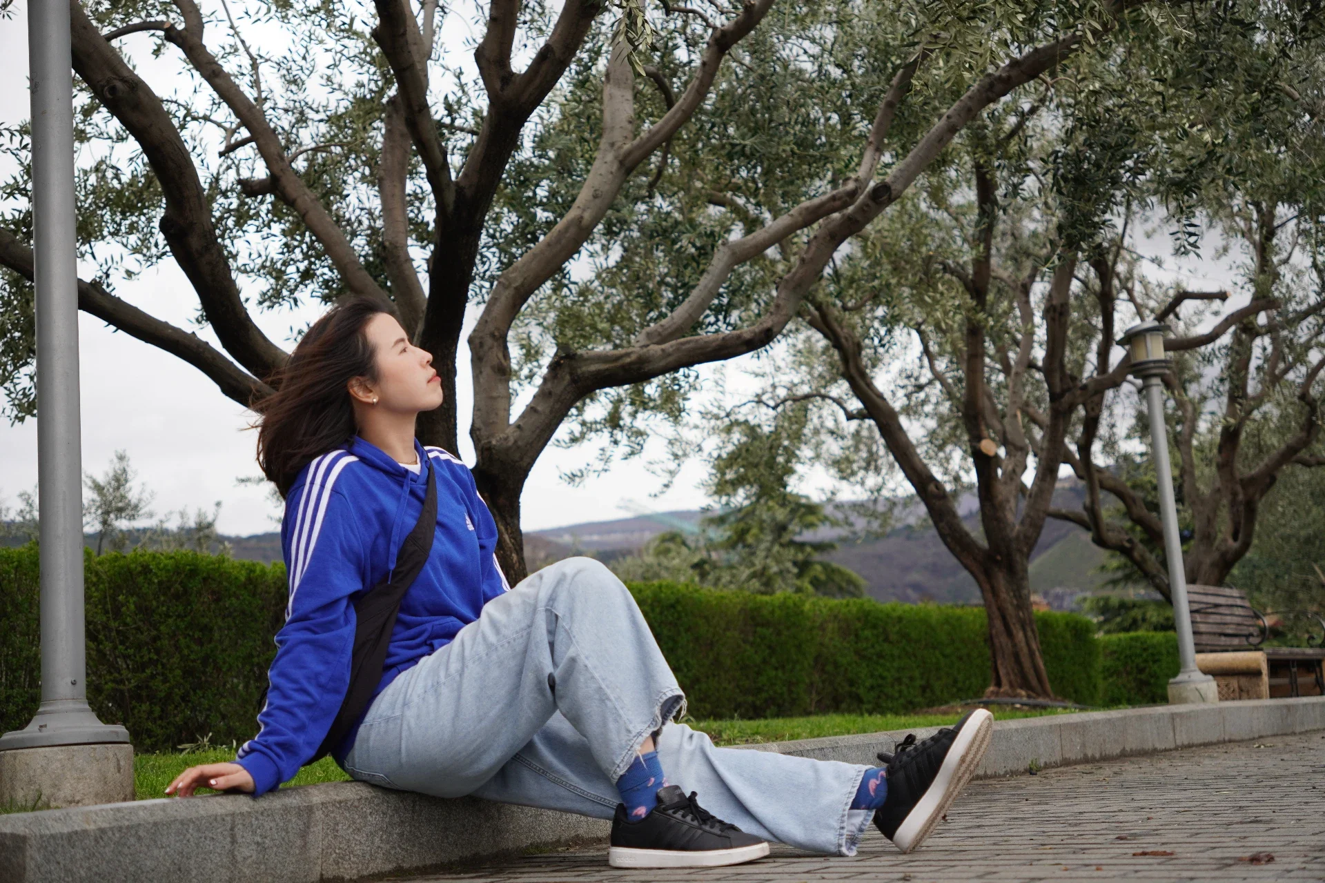 A young woman in a blue hoodie and light jeans sitting on a park bench with her legs stretched out, leaning back with her left hand resting on the ground, gazing up at the sky, surrounded by trees and greenery.
