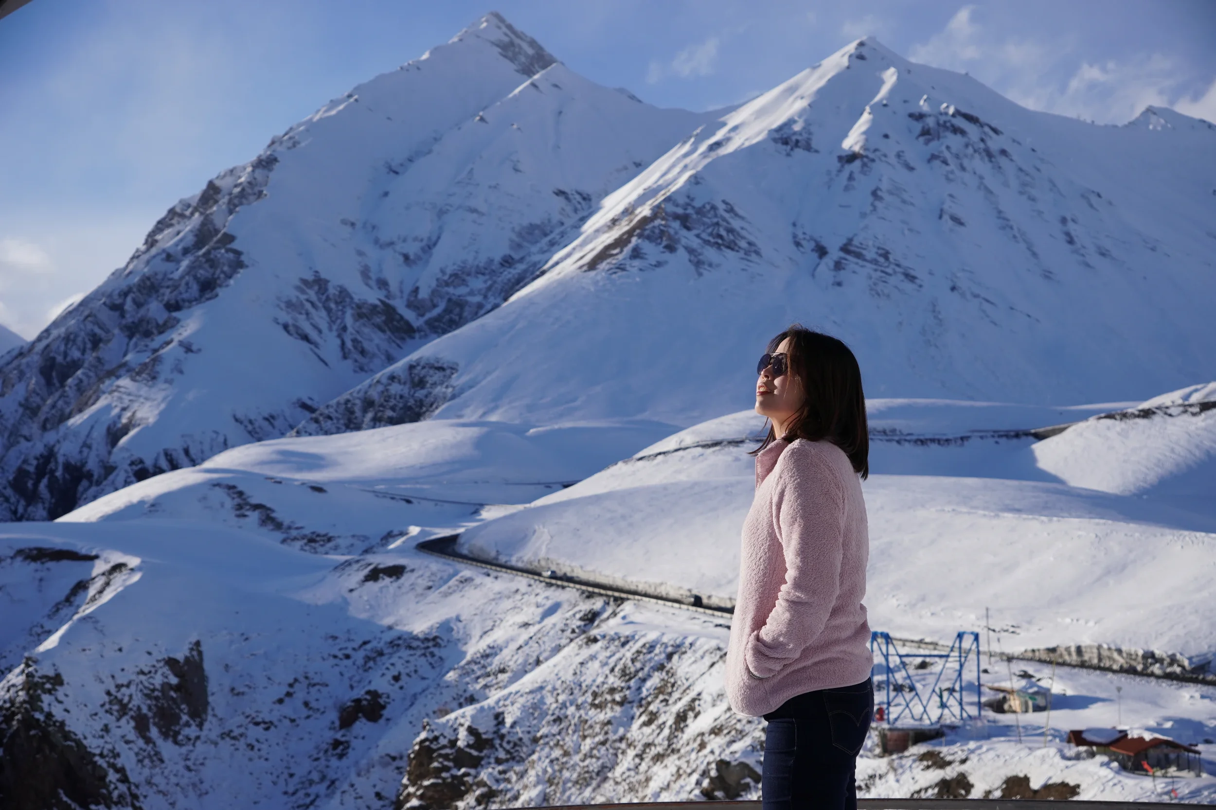 A woman with dark hair and sunglasses wearing a pink jacket stands in front of snow-covered mountains during daytime.