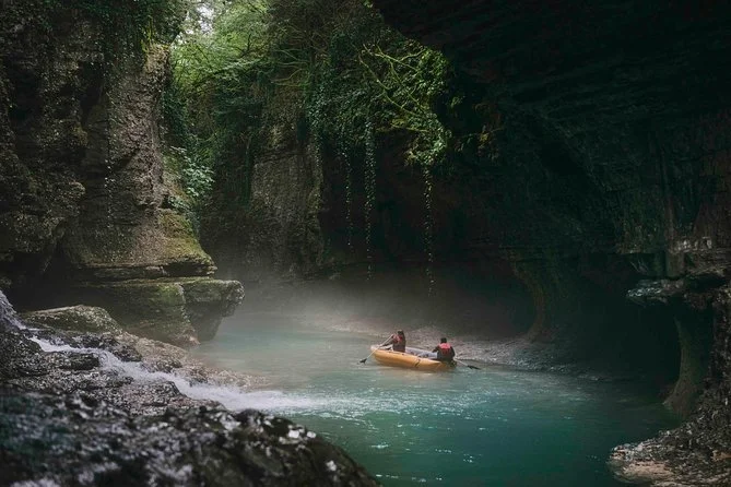 Two people kayaking in a narrow, rocky canyon with lush green trees overhead and a misty atmosphere.