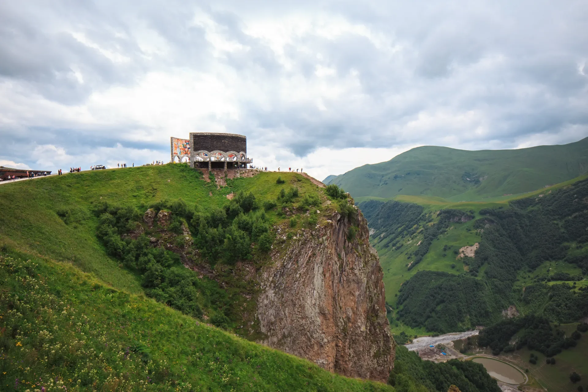 A scenic view of a high green hill with a modern, artistic building at the top, overlooking a lush valley and distant hills under a cloudy sky.