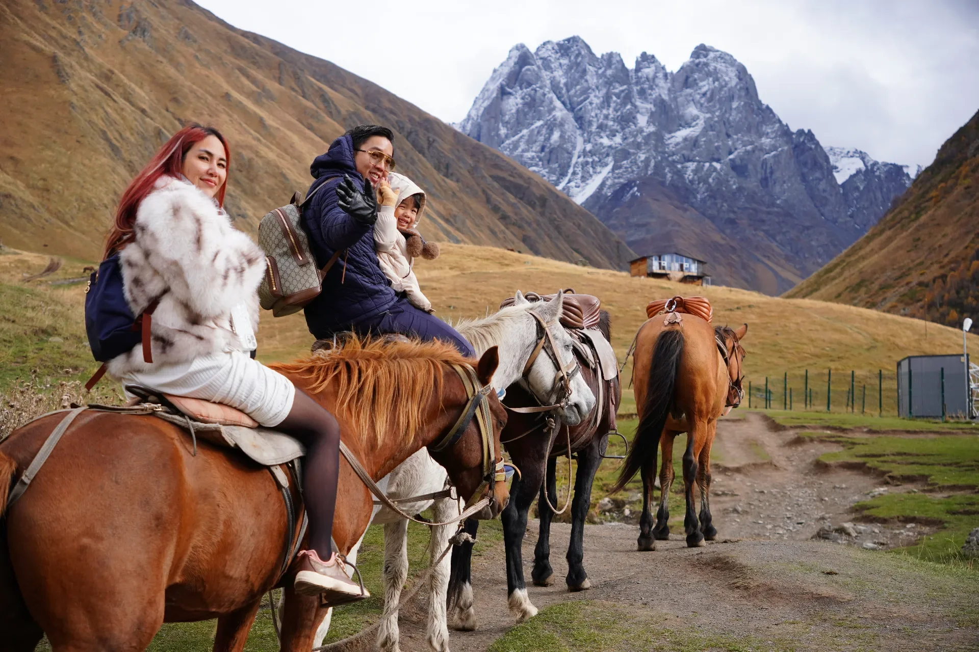 Three women and a child riding horses in a mountainous landscape with snow-capped peaks in the background.