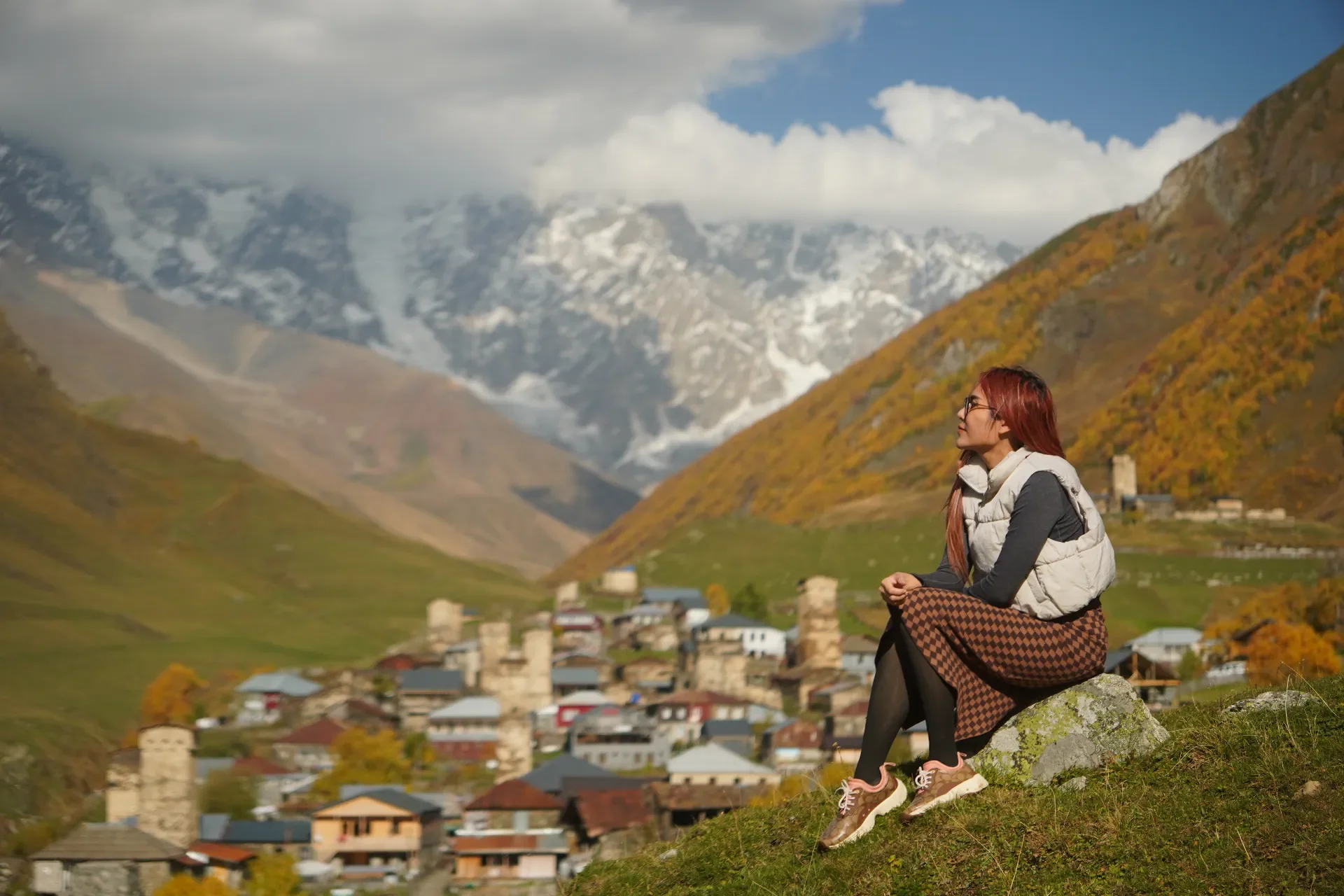 A woman with red hair, glasses, and a gray vest sitting on a rock on a hillside, overlooking a mountain village with a backdrop of snow-capped mountains and a cloudy sky.