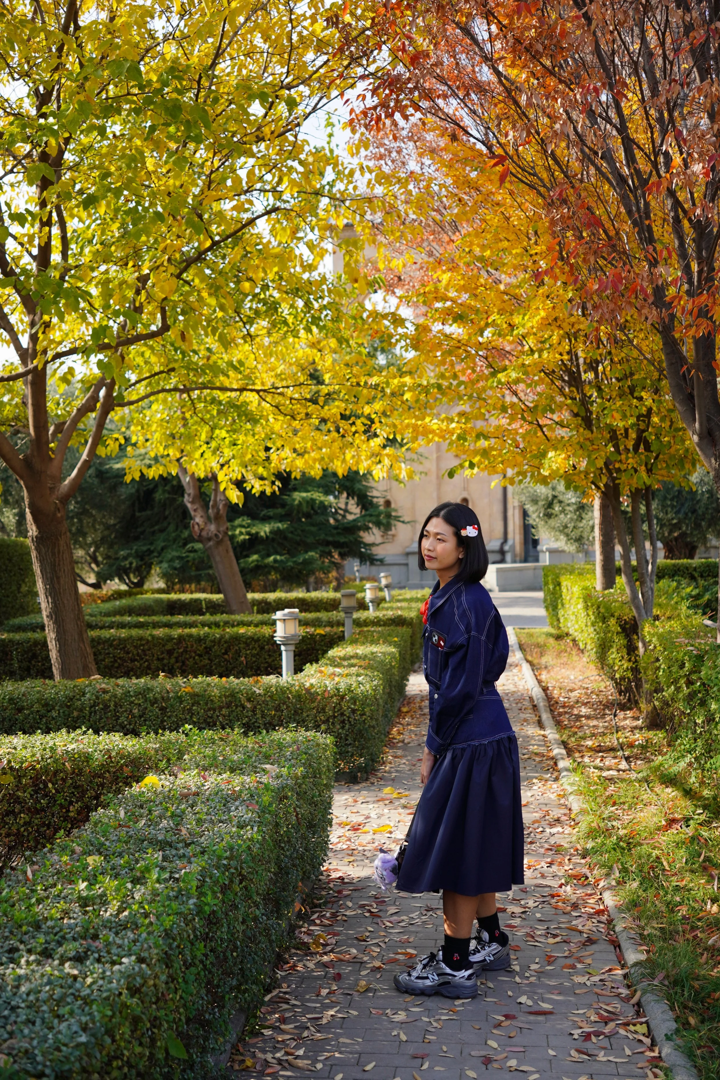 A young woman standing on a brick walkway lined with neatly trimmed bushes and colorful autumn trees, wearing a navy blue jacket and skirt, holding a plush toy, and looking into the distance.