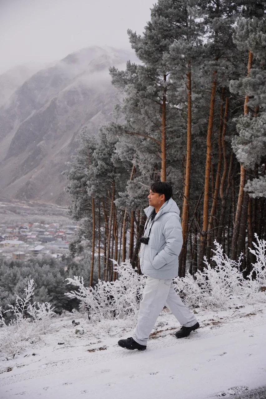 A man dressed in a white winter coat and pants walking along a snow-covered path in a forest with snow-covered trees, mountains in the background, and foggy weather.