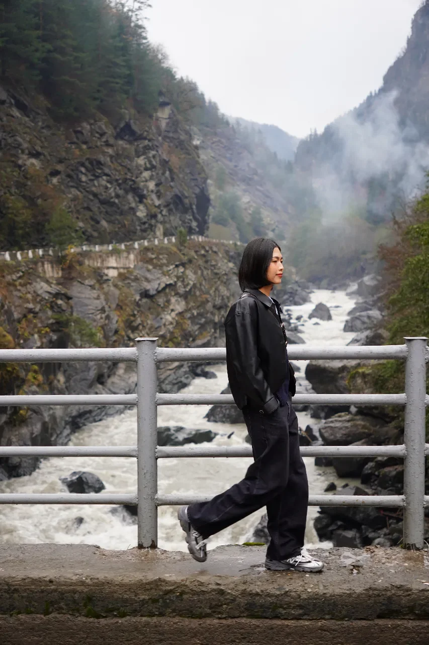 A woman standing on a bridge near a river with rocks, surrounded by mountainous terrain with fog or smoke, wearing a black jacket, black pants, and sneakers.
