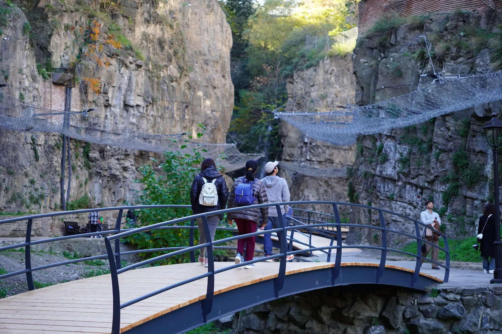 People walking on a curved wooden bridge in a canyon with high rock walls, some with waterfalls and greenery, and suspension bridges overhead.