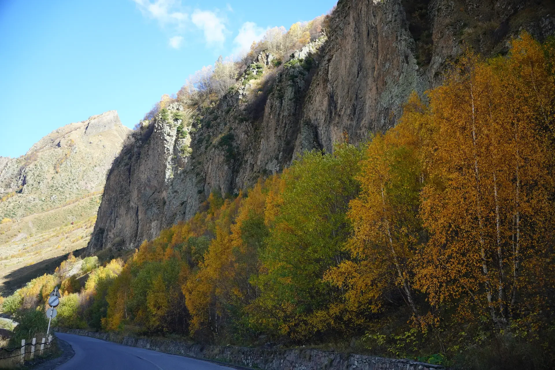 A winding mountain road surrounded by colorful autumn trees and towering rocky cliffs under a blue sky.