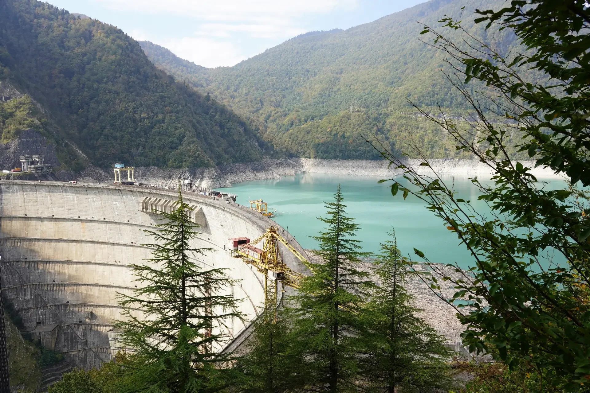 A large dam with a reservoir surrounded by green mountains, with trees in the foreground and a partly cloudy sky.