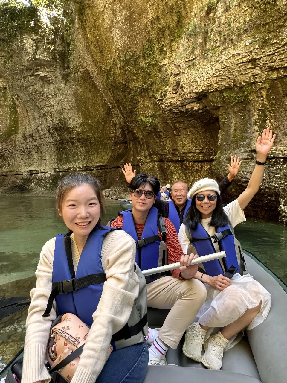 Four people, three women and one man, in a boat on a river, wearing life jackets, with a rocky cliffside in the background, enjoying a rafting adventure.