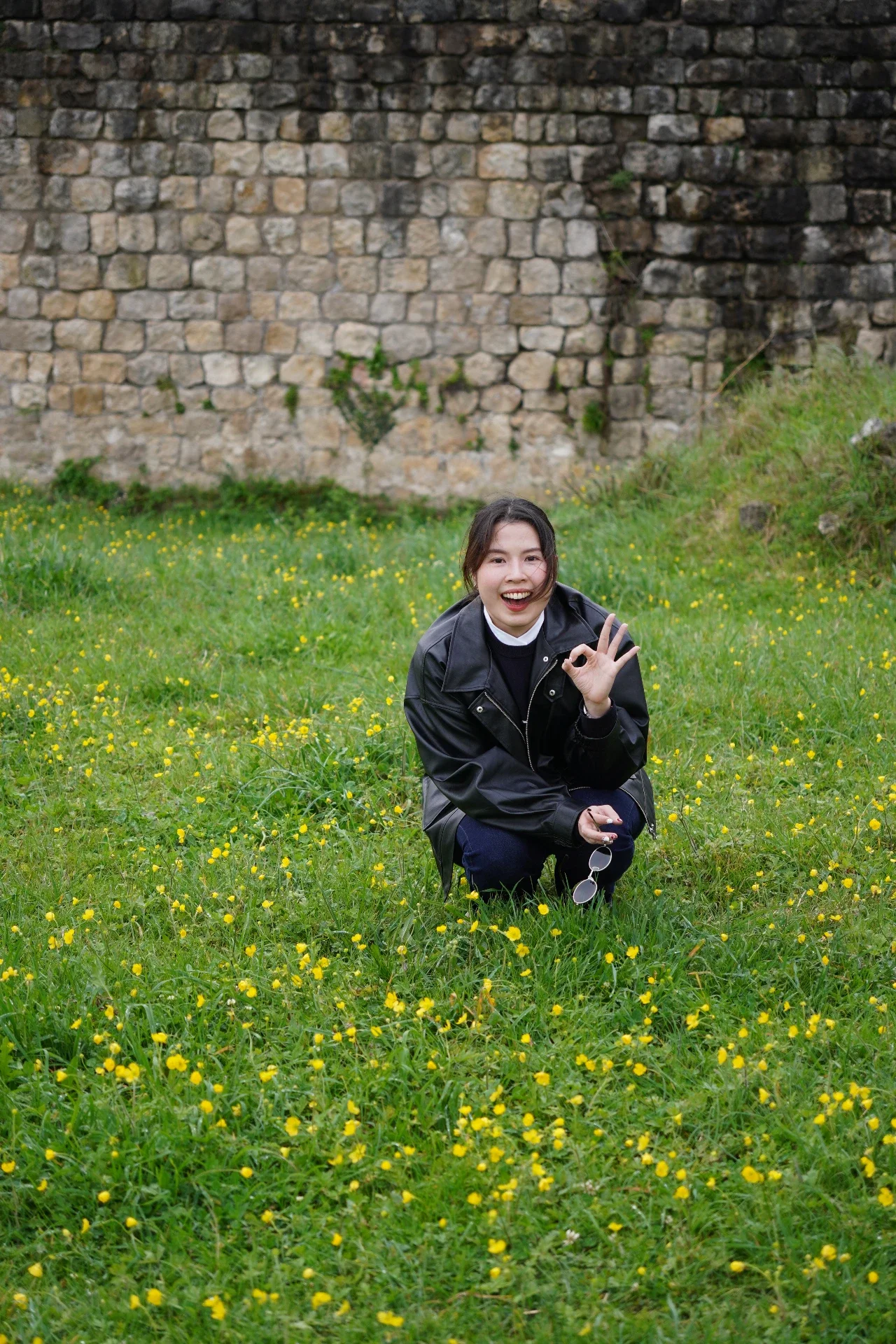 Smiling woman with dark hair, wearing a black jacket, squatting on a grassy field with small yellow flowers, holding sunglasses, and making an OK gesture with her right hand, in front of a stone wall.