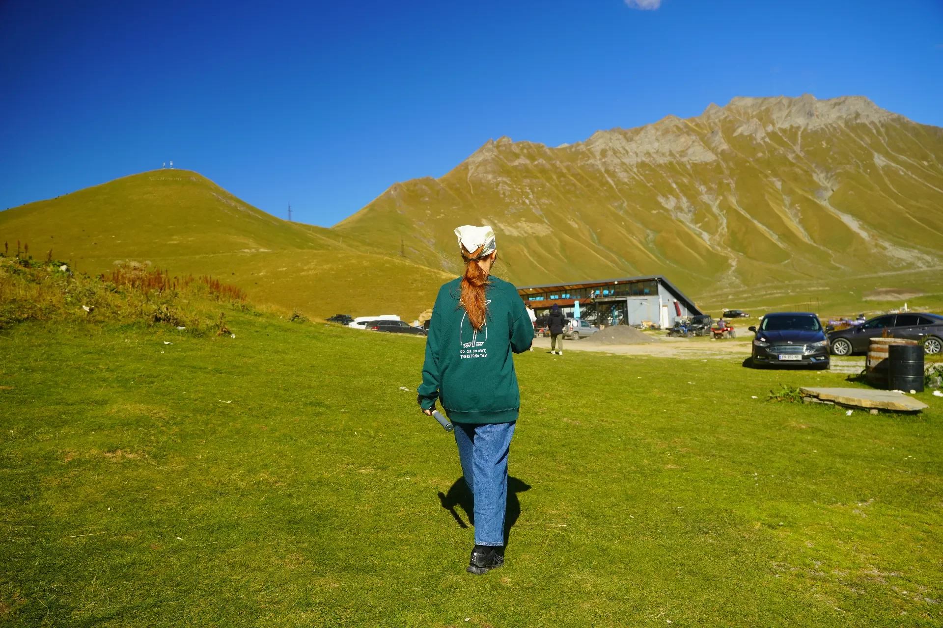 A person with long hair in a ponytail, wearing a green hoodie, blue jeans, a beige headscarf, and black shoes, walking on a grassy field towards a building at the foot of mountains under a clear blue sky.