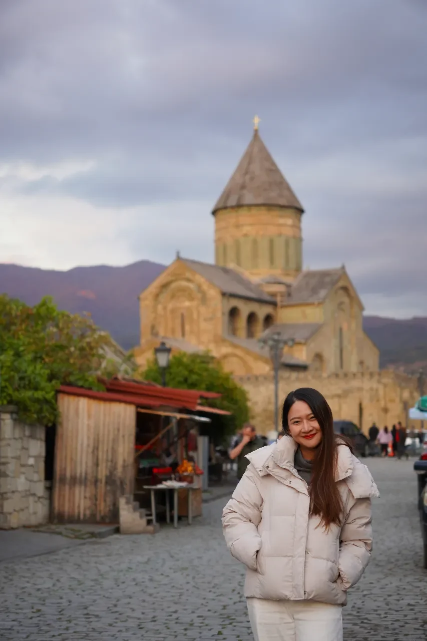 A smiling woman with long dark hair in a white winter coat stands on a cobblestone street in front of an old stone church with a tall bell tower, under a cloudy sky.