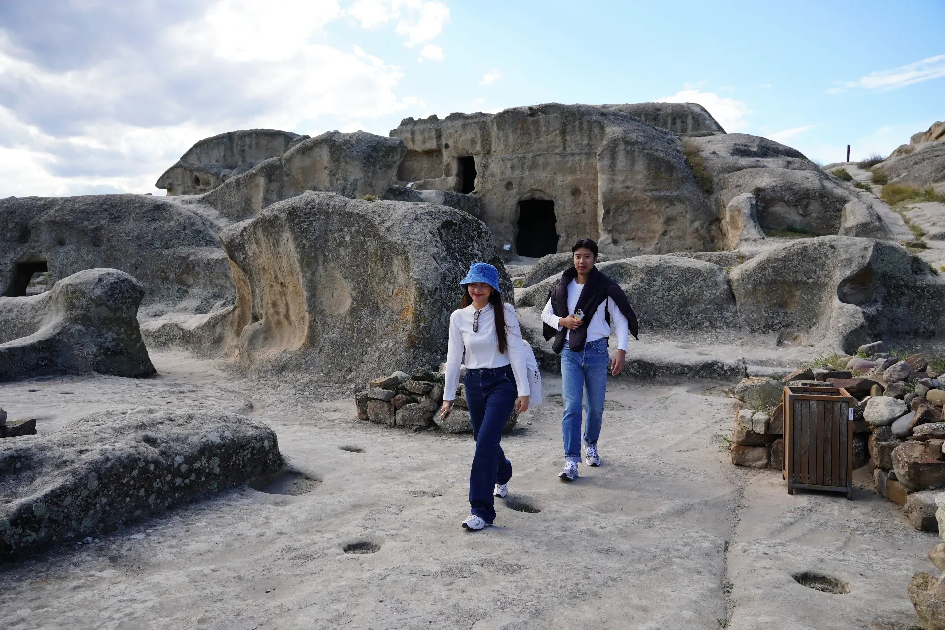 Two women walk along a rocky path with ancient rock-cut dwellings behind them, under a partly cloudy sky.
