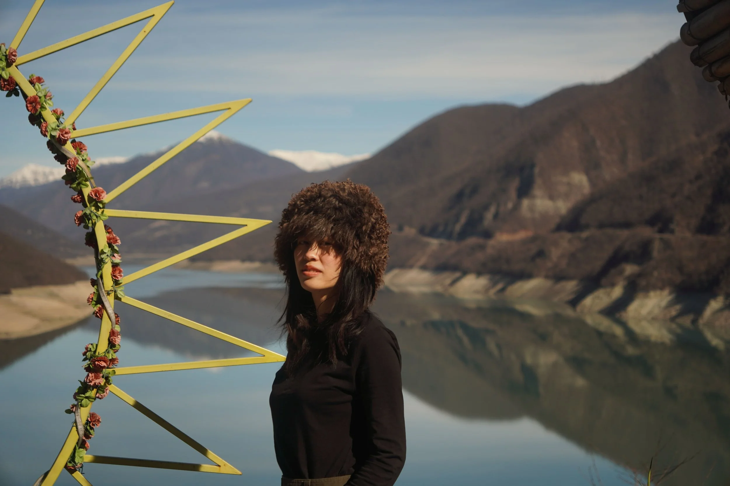 A woman with dark hair wearing a fuzzy brown hat and a black top, standing outdoors near a reflective lake and mountains, with a decorative yellow metal structure with pink flowers on it to her left.