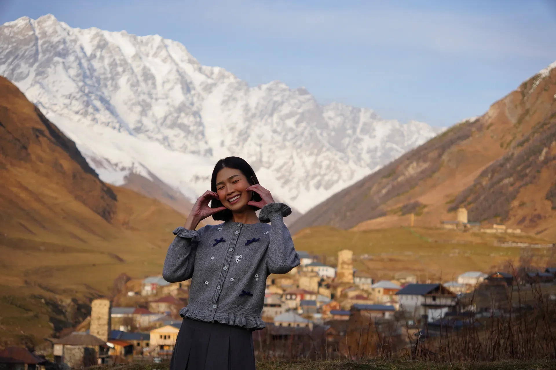 A woman smiling and making a heart shape with her hands stands outdoors with a mountainous landscape in the background, including snow-capped peaks and a village on the hillside.