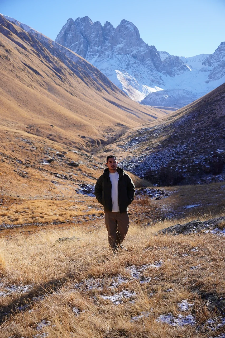 A man standing in a mountain valley with snow-capped peaks in the background, wearing a dark jacket, white shirt, and brown pants.