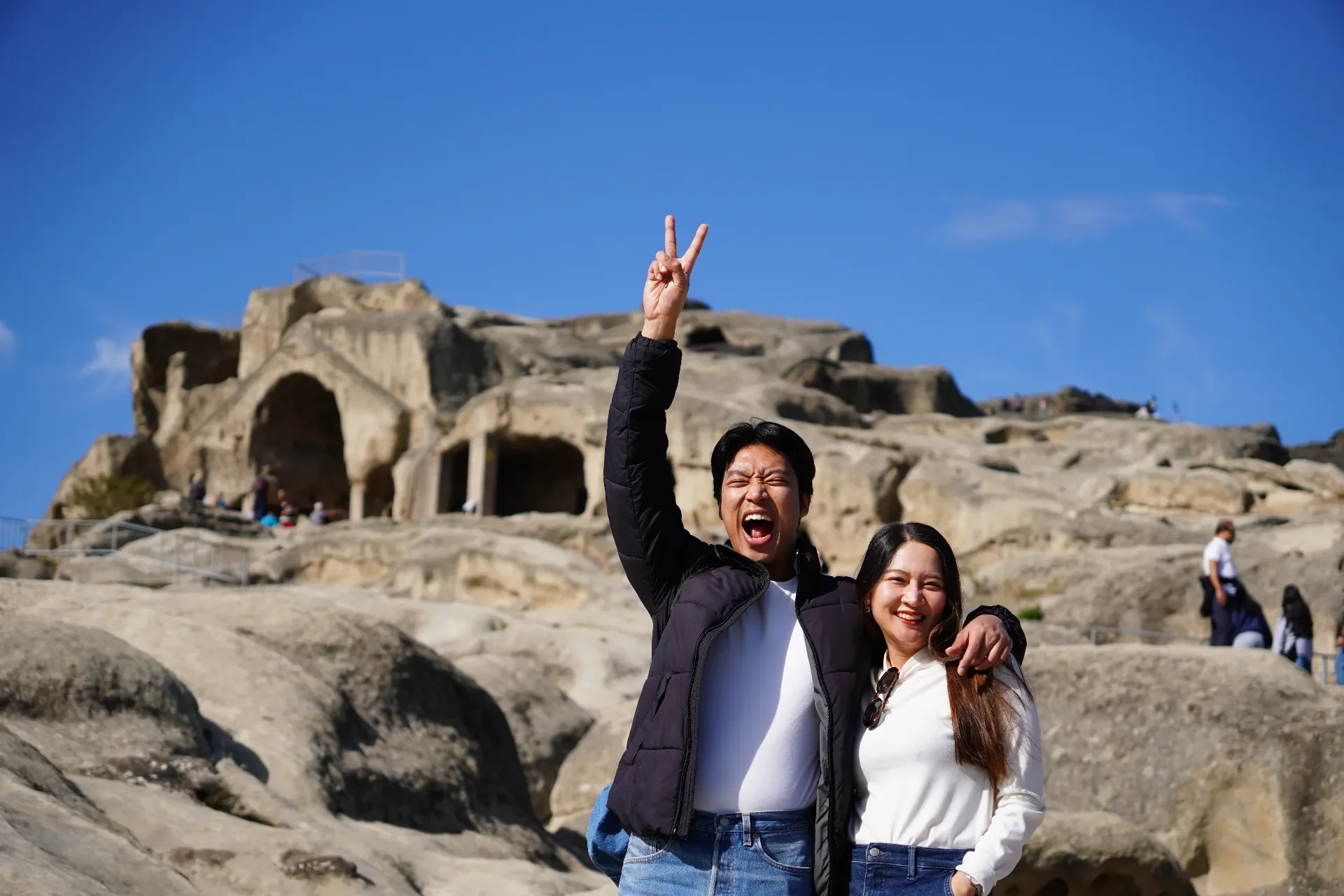 Two people smiling and posing with rock formations in the background, one making a peace sign with hand raised, under a blue sky.