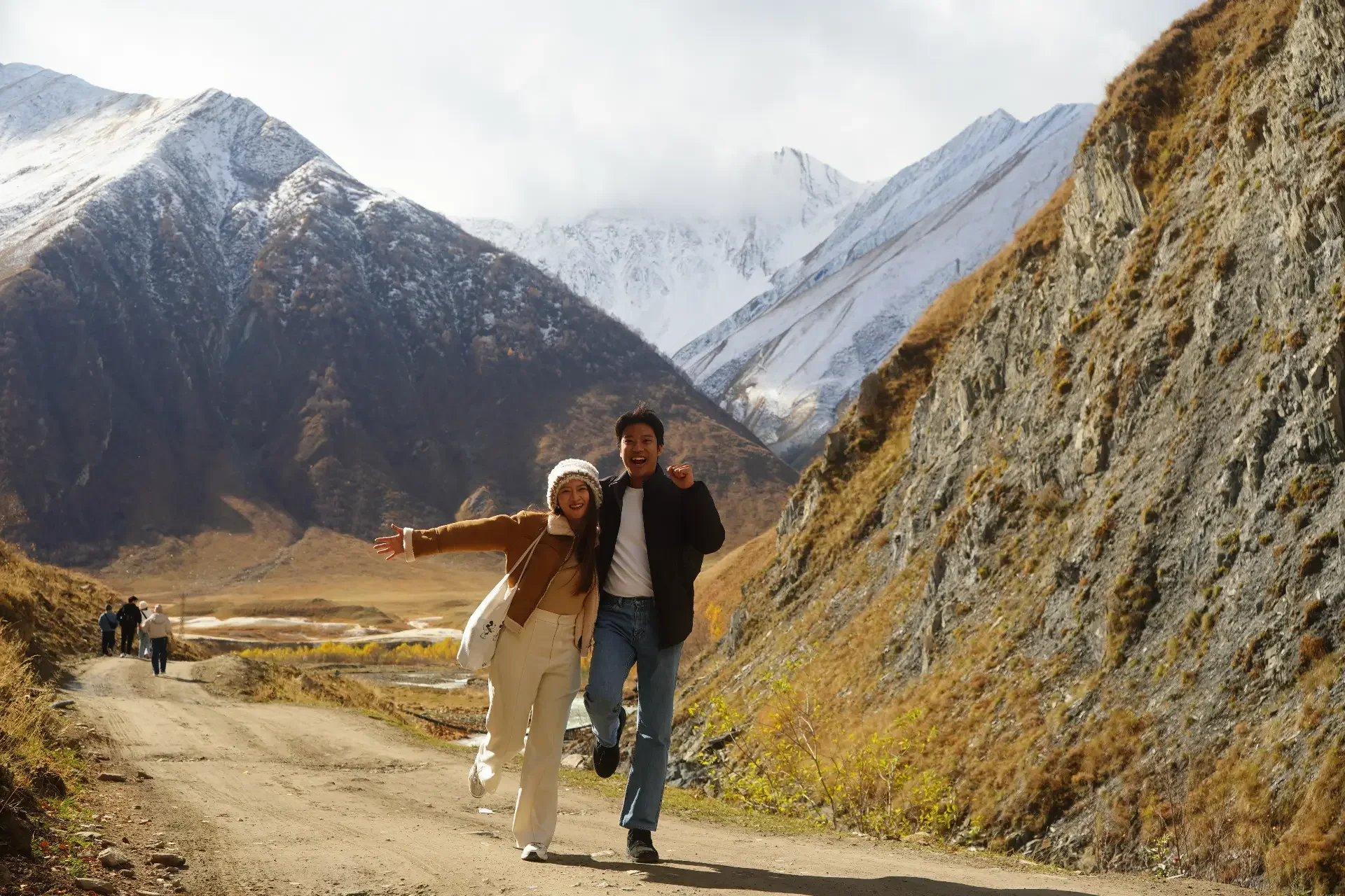 Two friends hiking on a dirt trail in a mountain landscape with snow-capped peaks, dry grass, and rocky slopes, smiling and enjoying the outdoors.