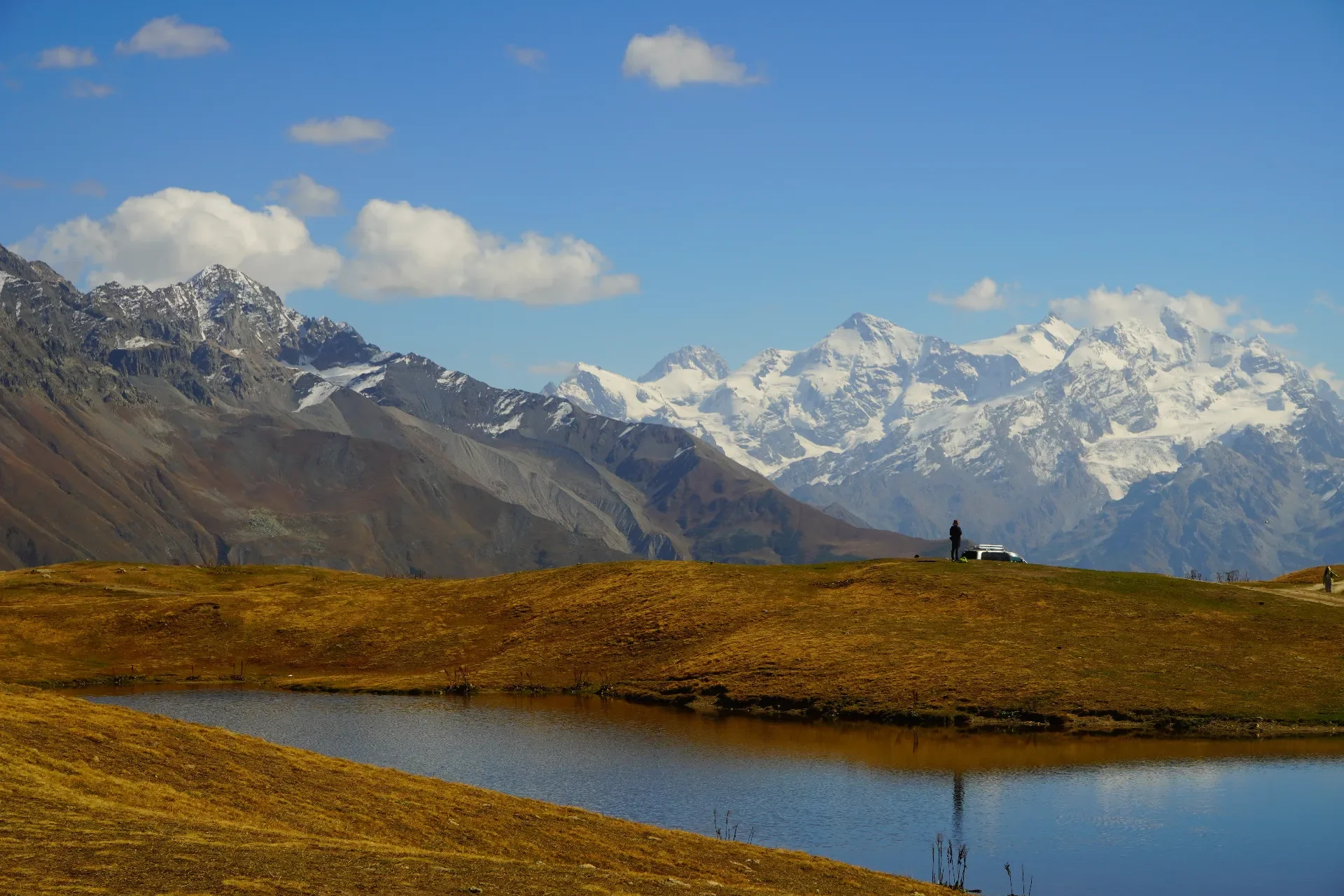 A mountain landscape with snow-capped peaks in the background, rolling brown hills in the foreground, a small pond, and a few people near a bench and cars.