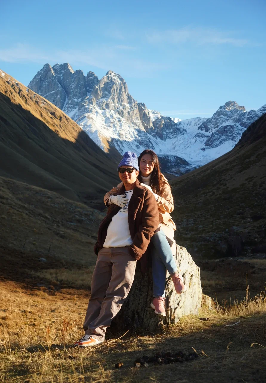Two women outdoors in a mountainous landscape with snow-capped peaks in the background. One woman is standing, wearing sunglasses, a purple beanie, and a brown jacket, while the other woman is sitting on a large rock, smiling, with long hair, wearing