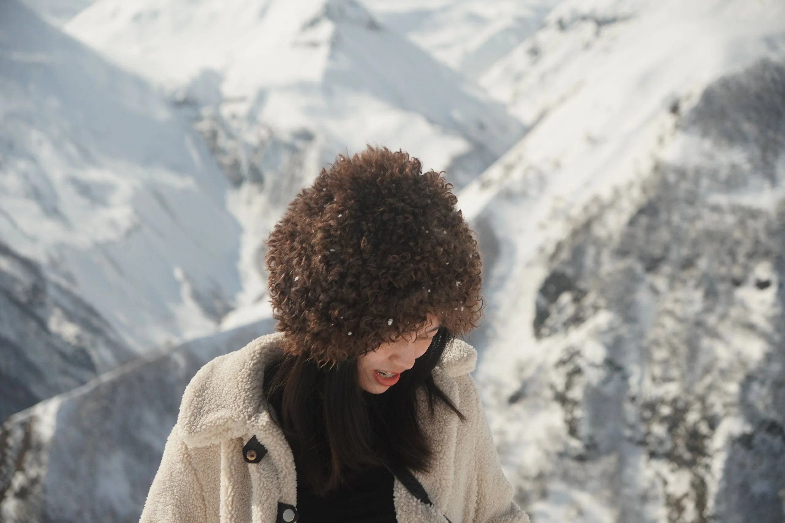 A woman with curly brown hair wearing a beige coat in a snowy mountain landscape