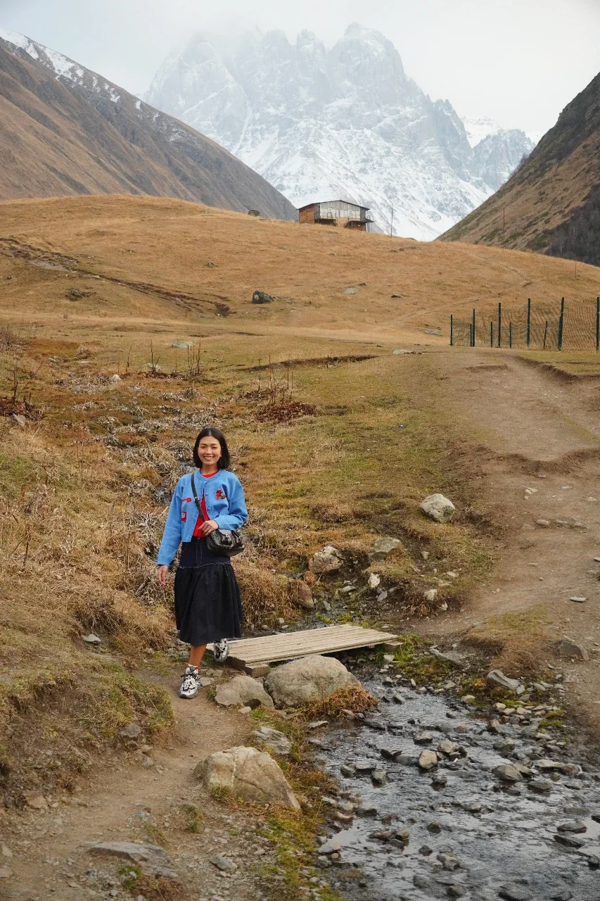 A woman standing on a small wooden bridge over a stream in a mountainous landscape with snow-capped peaks and a rustic house in the background.