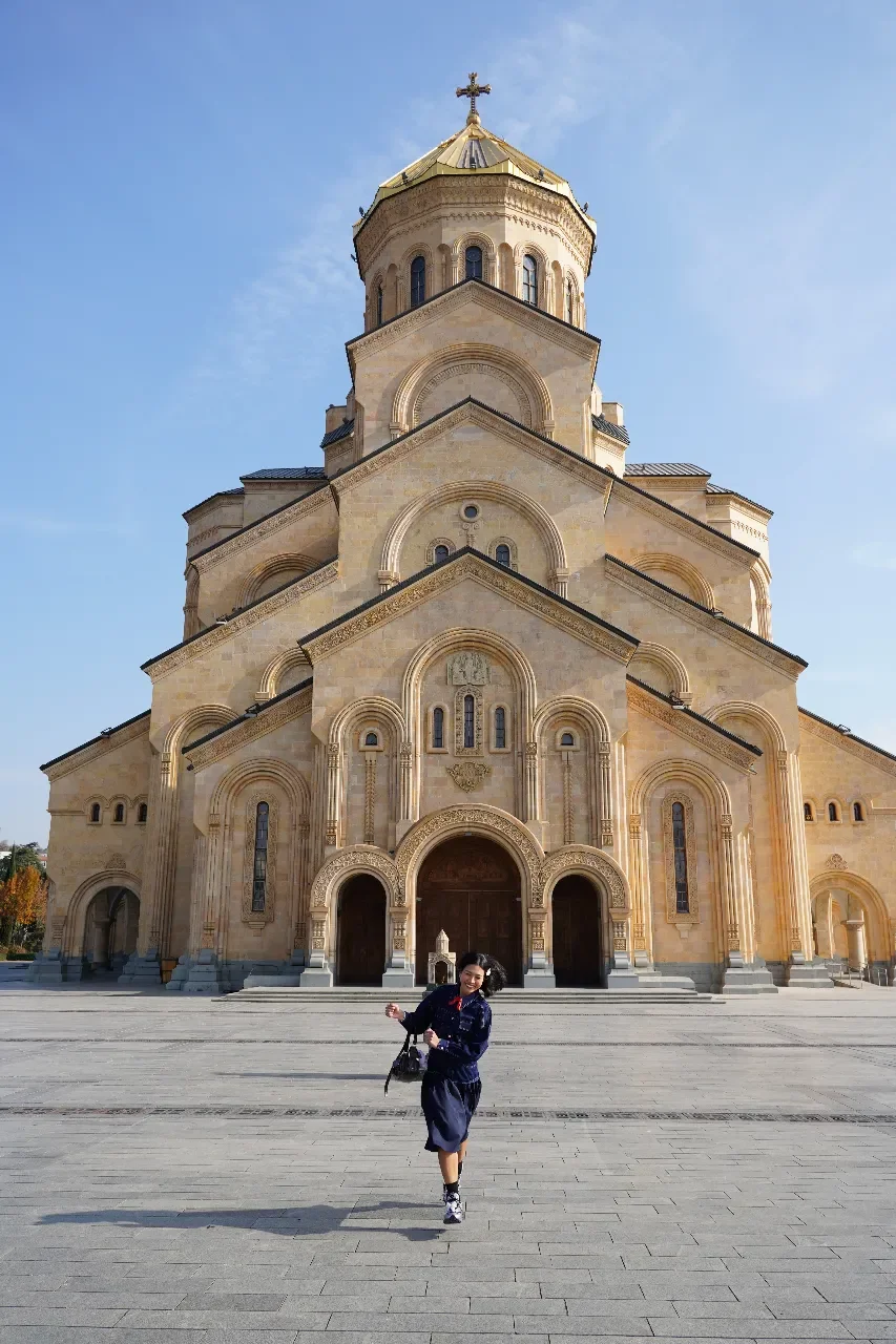 A woman in a navy outfit running in front of a large, ornate church with beige stone walls and a gold dome topped with a cross, under a clear blue sky.