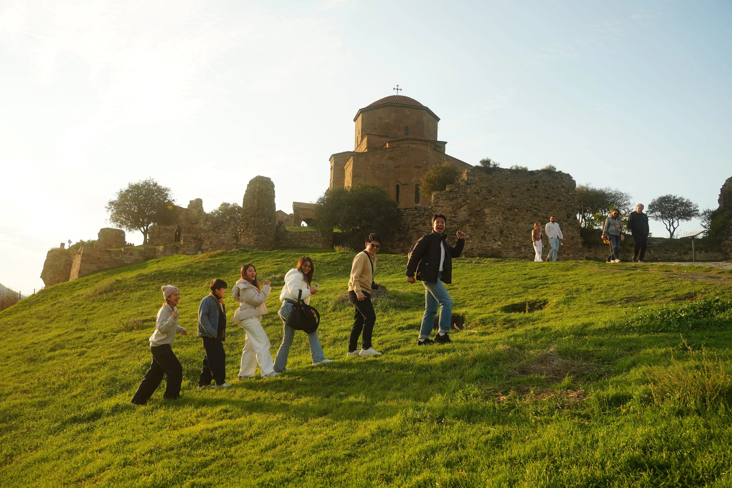 Group of people enjoying a walk up a grassy hill on a sunny day, with an ancient stone church and ruins in the background.
