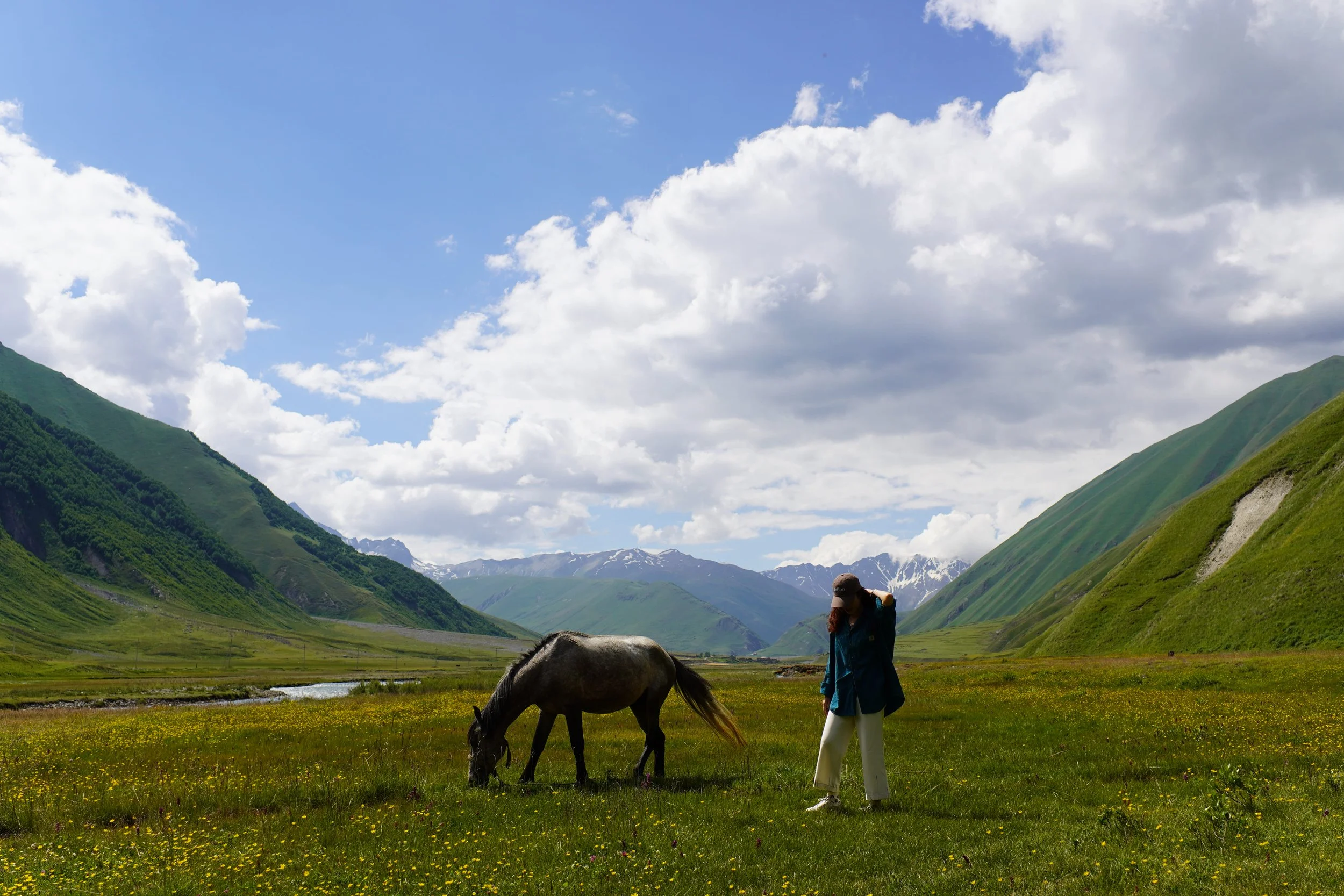 A person standing in a grassy valley with a horse grazing nearby, surrounded by green mountains and a partly cloudy sky.