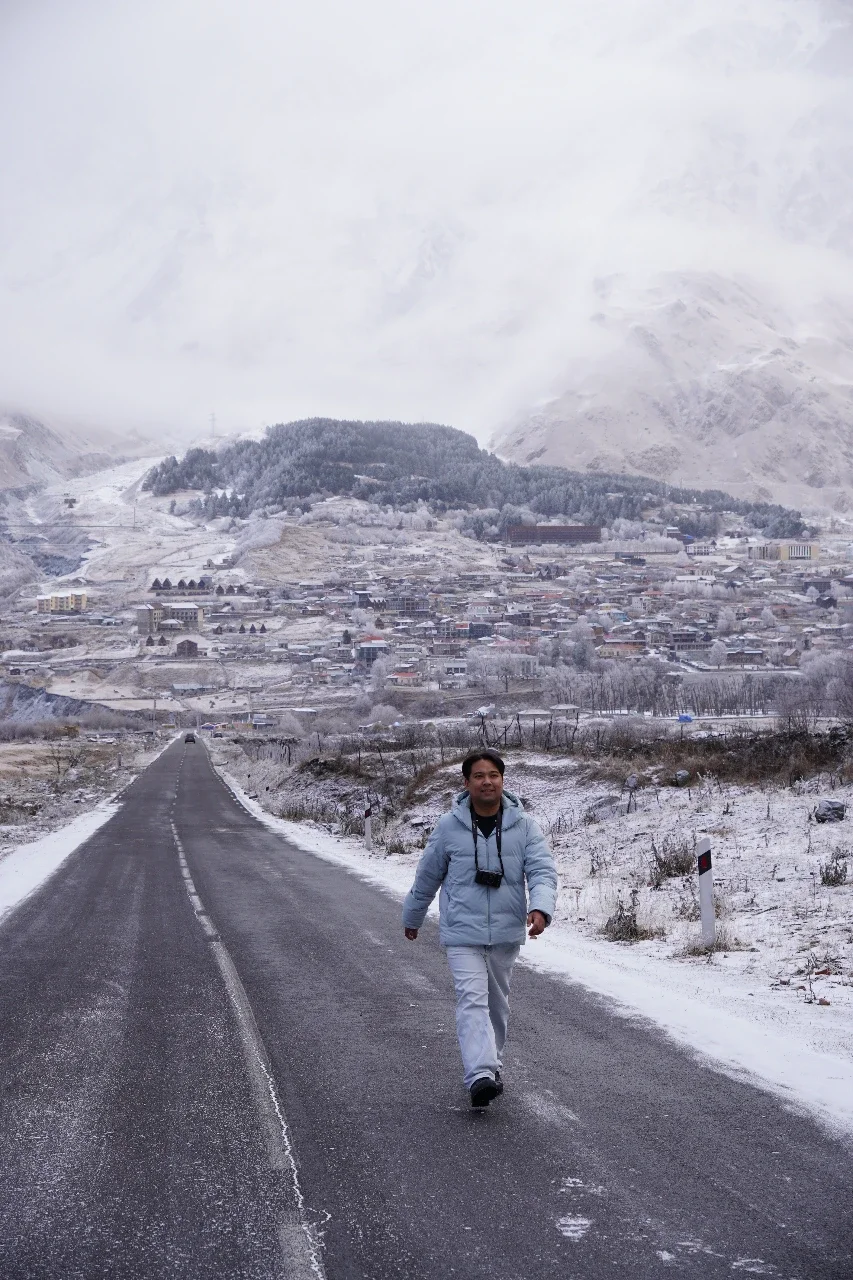 A man walking on a snow-covered road with a camera around his neck, against a backdrop of a snowy town and mountains.