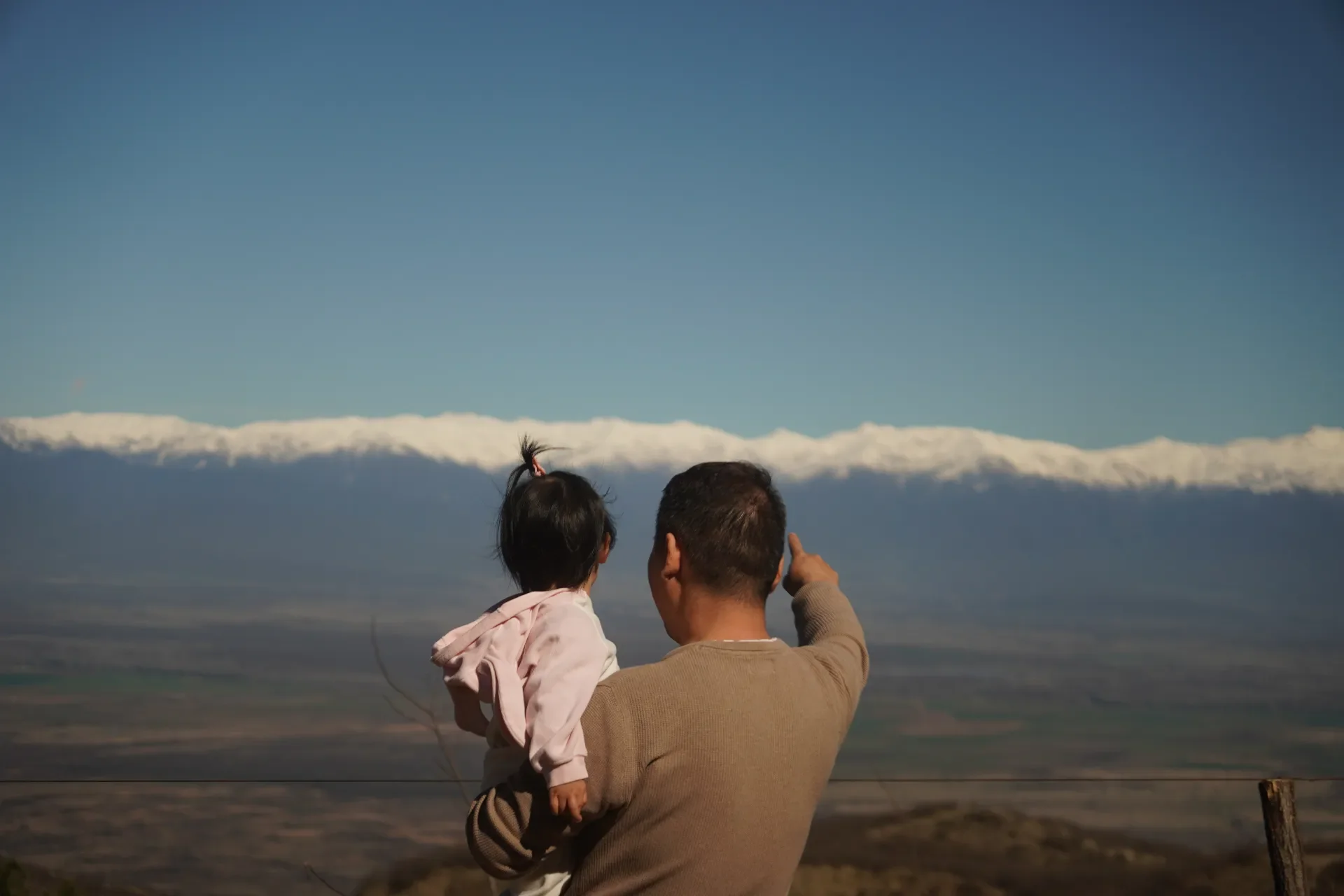 A man holding a child and pointing towards a snow-capped mountain in the distance under a clear blue sky.