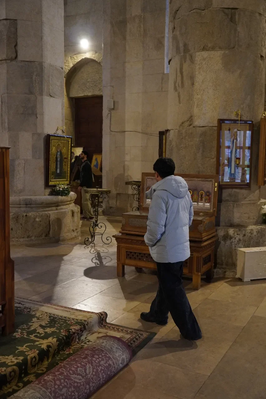 A person in a light gray jacket inside a church, looking at religious icons displayed in wooden frames on stands. The church has stone walls and a tiled floor, with a patterned rug on the floor.
