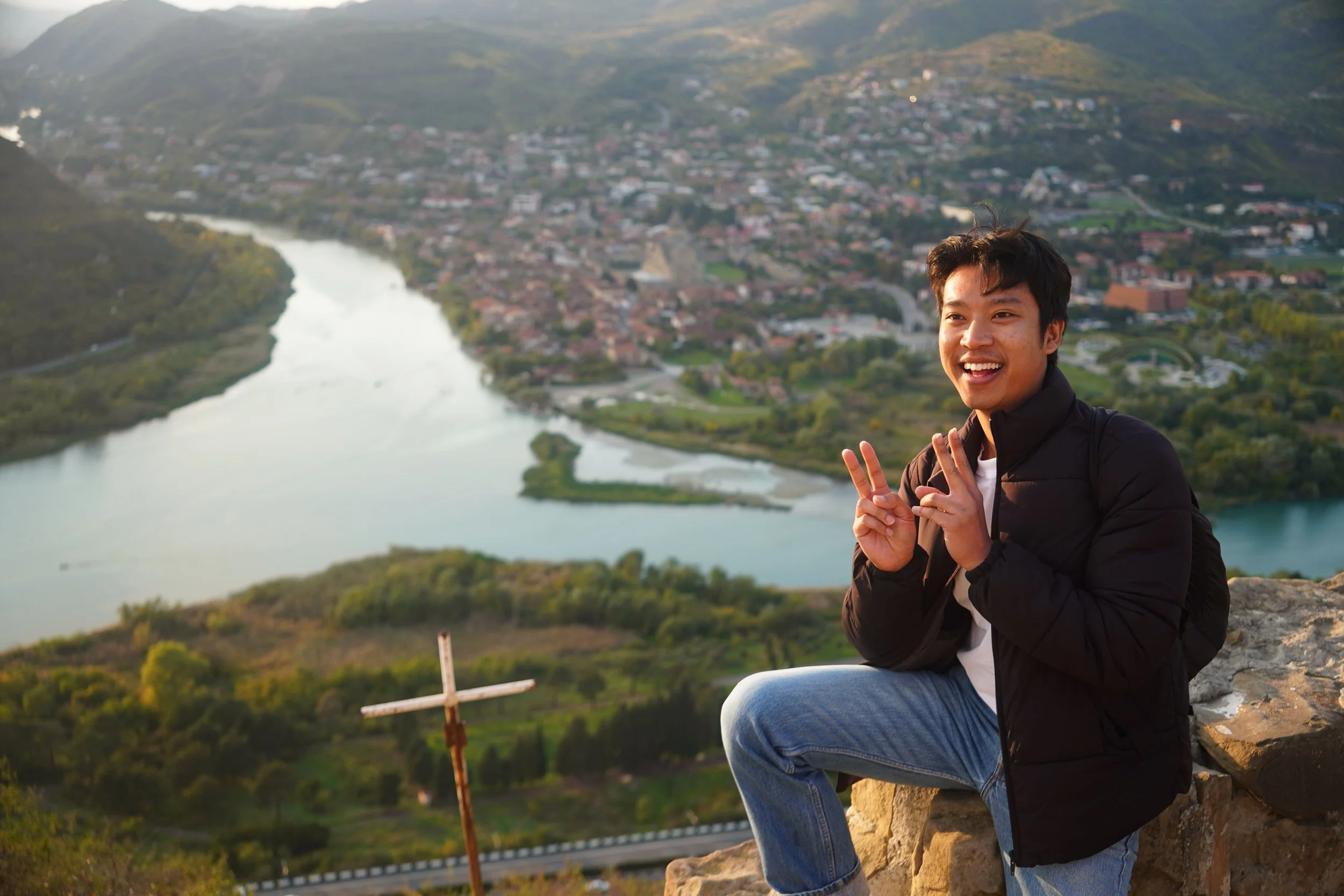 A young man sitting on a rock with a wide smile, making peace signs with both hands, overlooking a river and a town below from a hilltop during sunset.