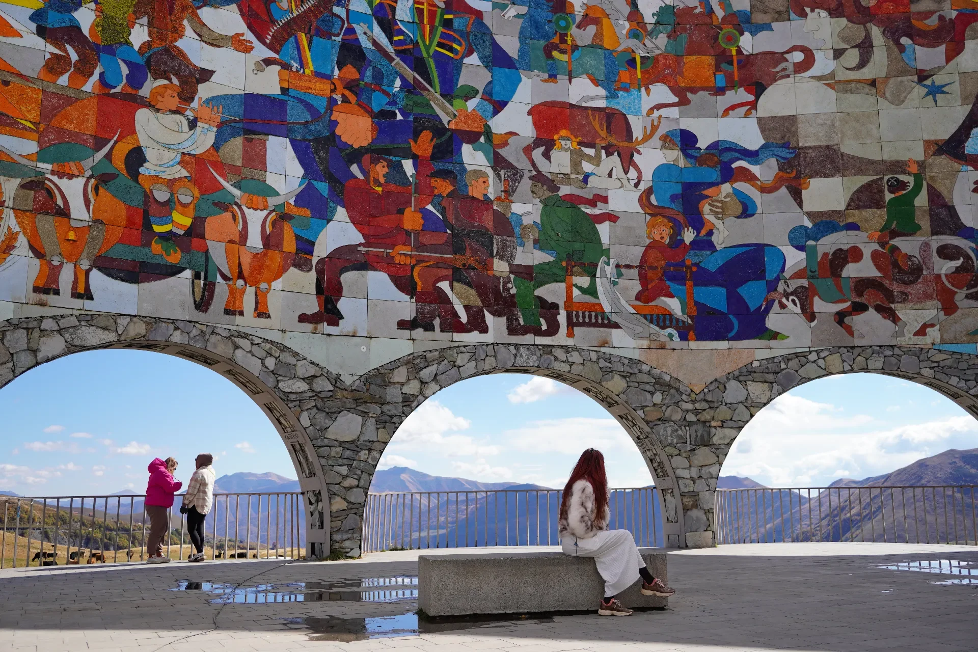 A person with red hair and white clothing sitting on a stone bench, looking out at mountains through three archways. On the left, two people are standing and talking, with one in a pink jacket. The mural above features colorful, stylized scenes with 