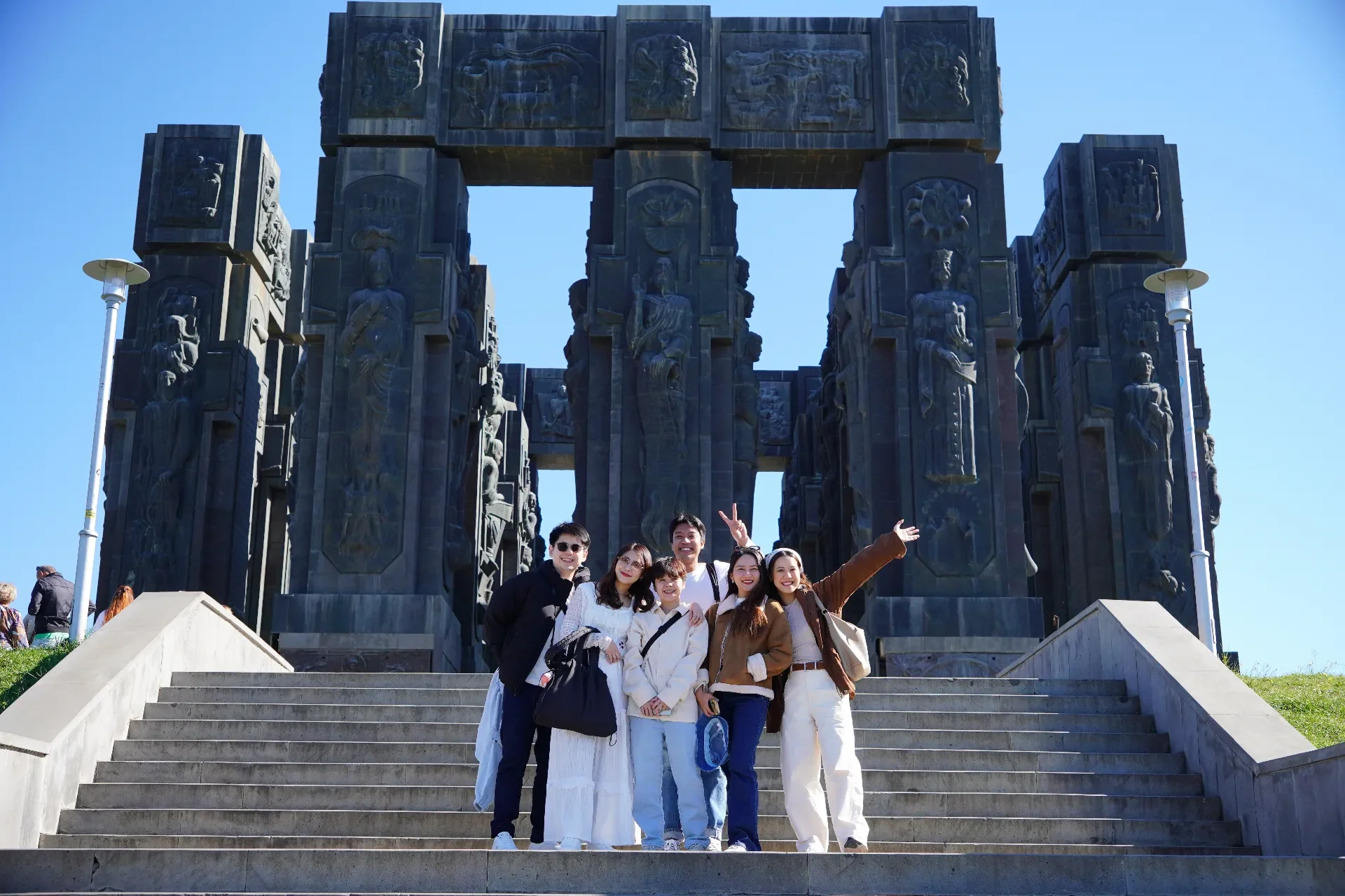 Five young adults standing on steps in front of a large, dark, sculpted archway with religious figures and symbols, under a clear blue sky.