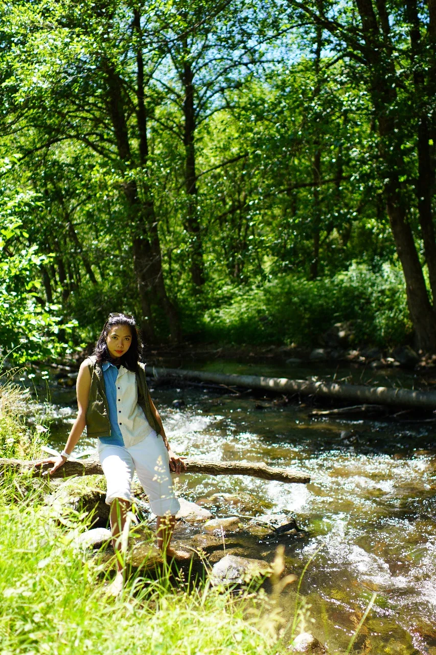 A woman standing by a flowing creek in a lush green forest on a sunny day.