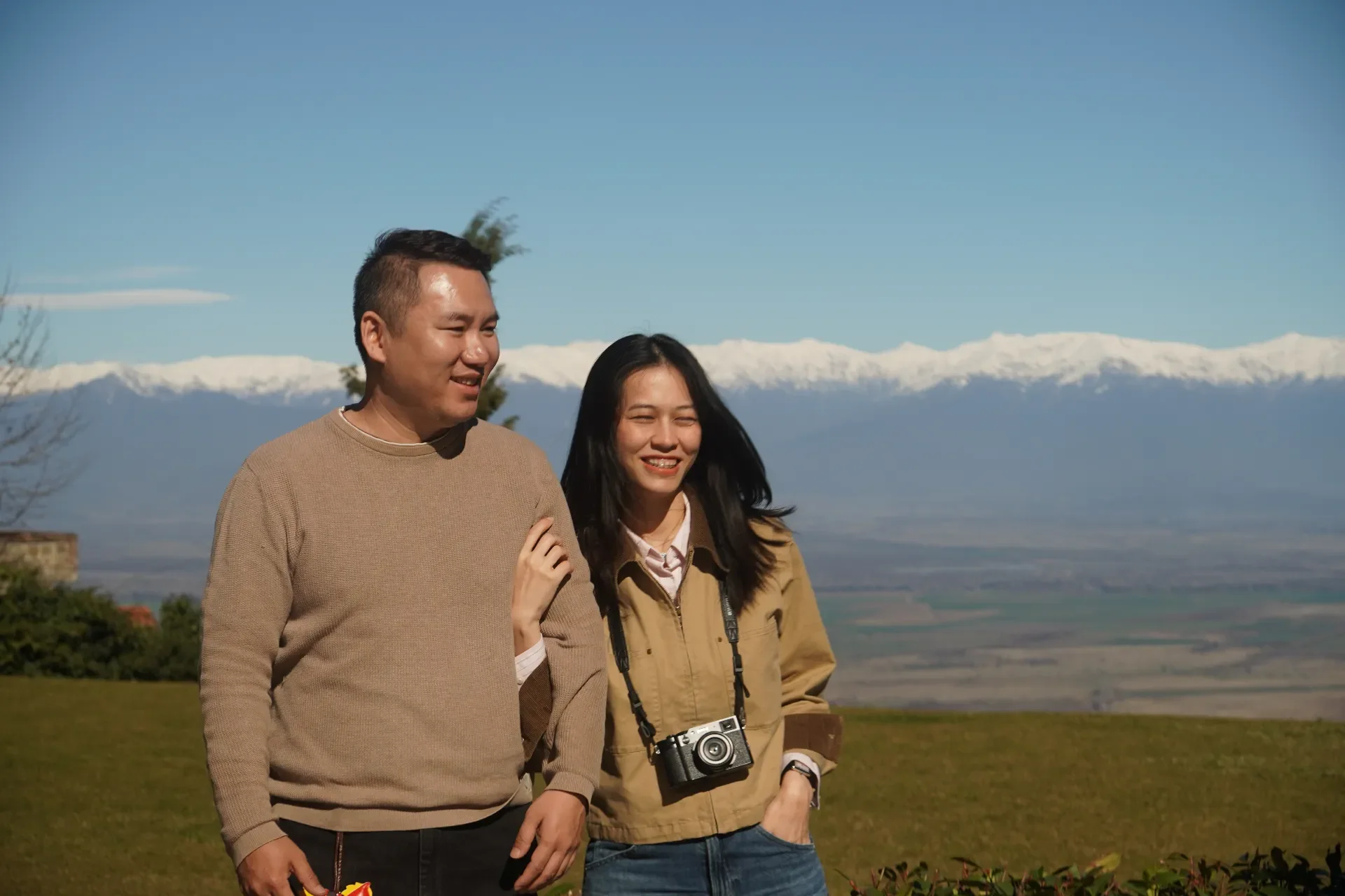 A man and a woman smiling and walking outdoors in front of snow-capped mountains. The woman has a camera hanging around her neck.