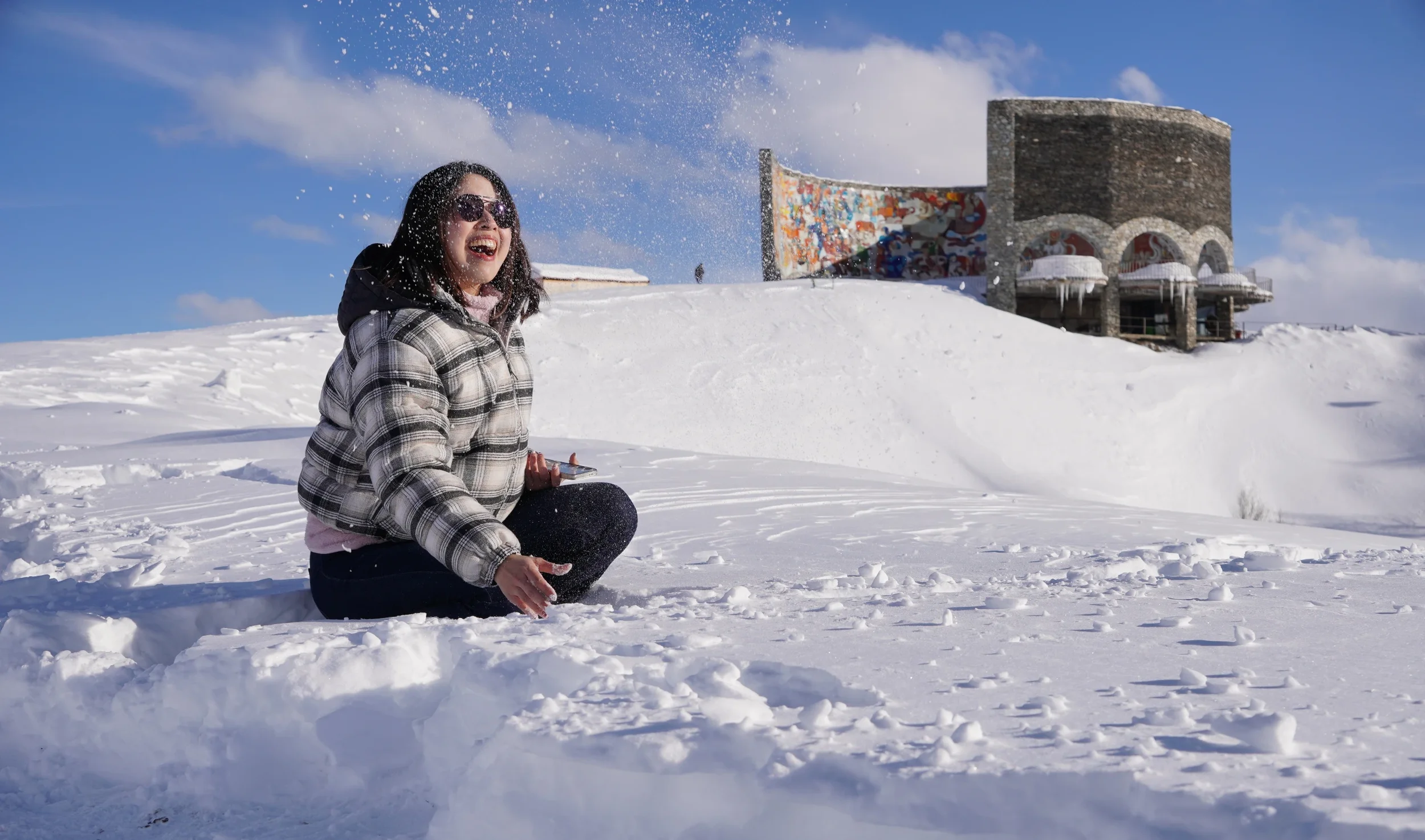 A woman sitting in snow, wearing sunglasses and a plaid jacket, smiling and playing with snow with a historic building and blue sky in the background.