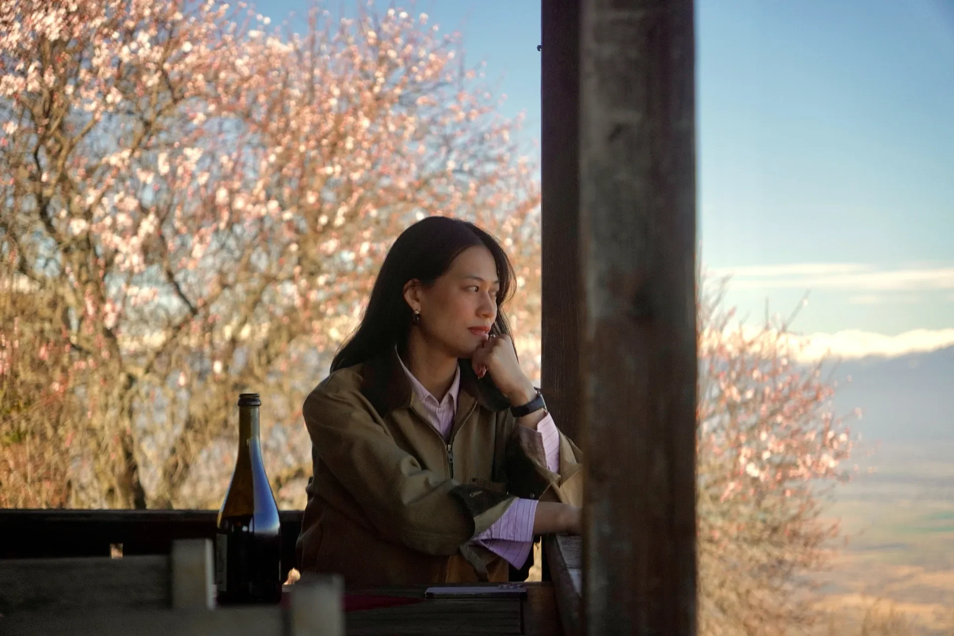 A woman with long dark hair is sitting at an outdoor location, resting her head on her hand, and looking thoughtfully into the distance. She is wearing a brown jacket over a pink striped shirt, with a bottle and some objects on the table in front of 