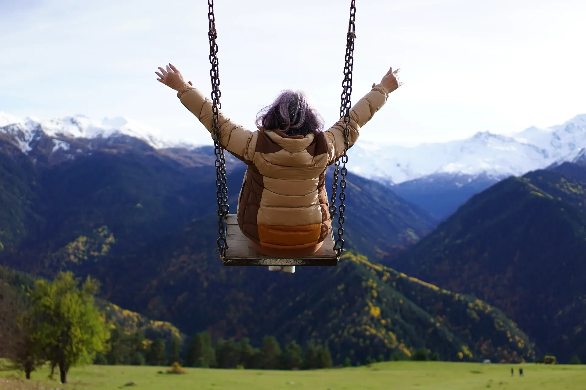 Person sitting on a swing with arms outstretched, overlooking a mountain landscape with snow-capped peaks and green valleys.