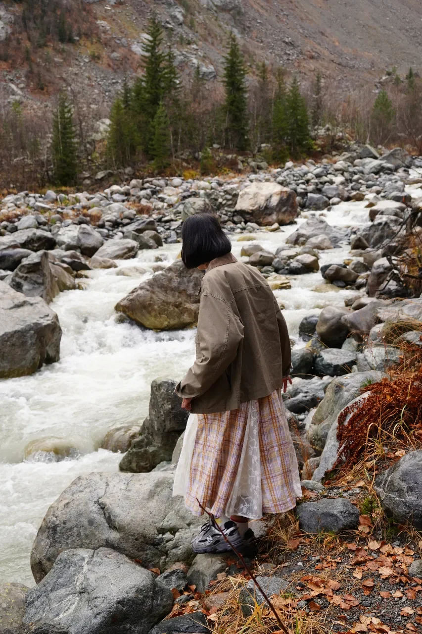 Person with short black hair wearing a tan jacket, plaid skirt, and sneakers standing on rocks beside a rushing mountain stream with a rocky hillside and pine trees in the background.