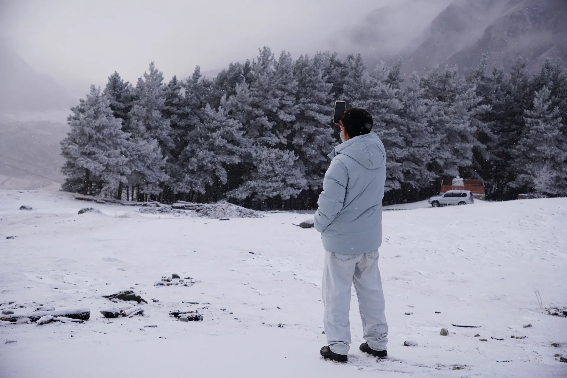 A person dressed in white winter clothes standing in a snowy landscape, taking a photo or recording a video of snow-covered trees with a smartphone.