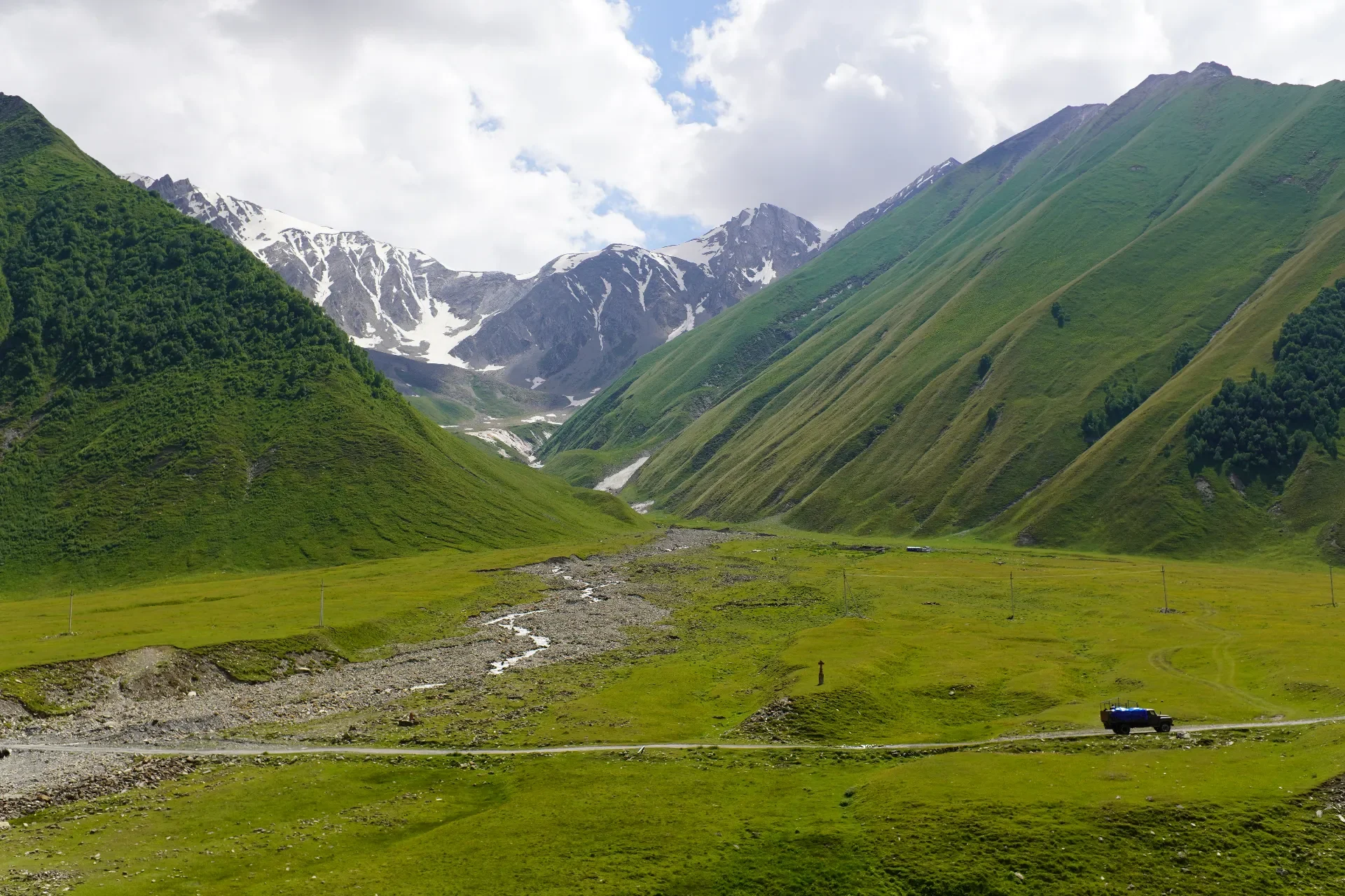 Green valley surrounded by mountains with snow-capped peaks in the background and a small vehicle on a dirt road in the foreground.