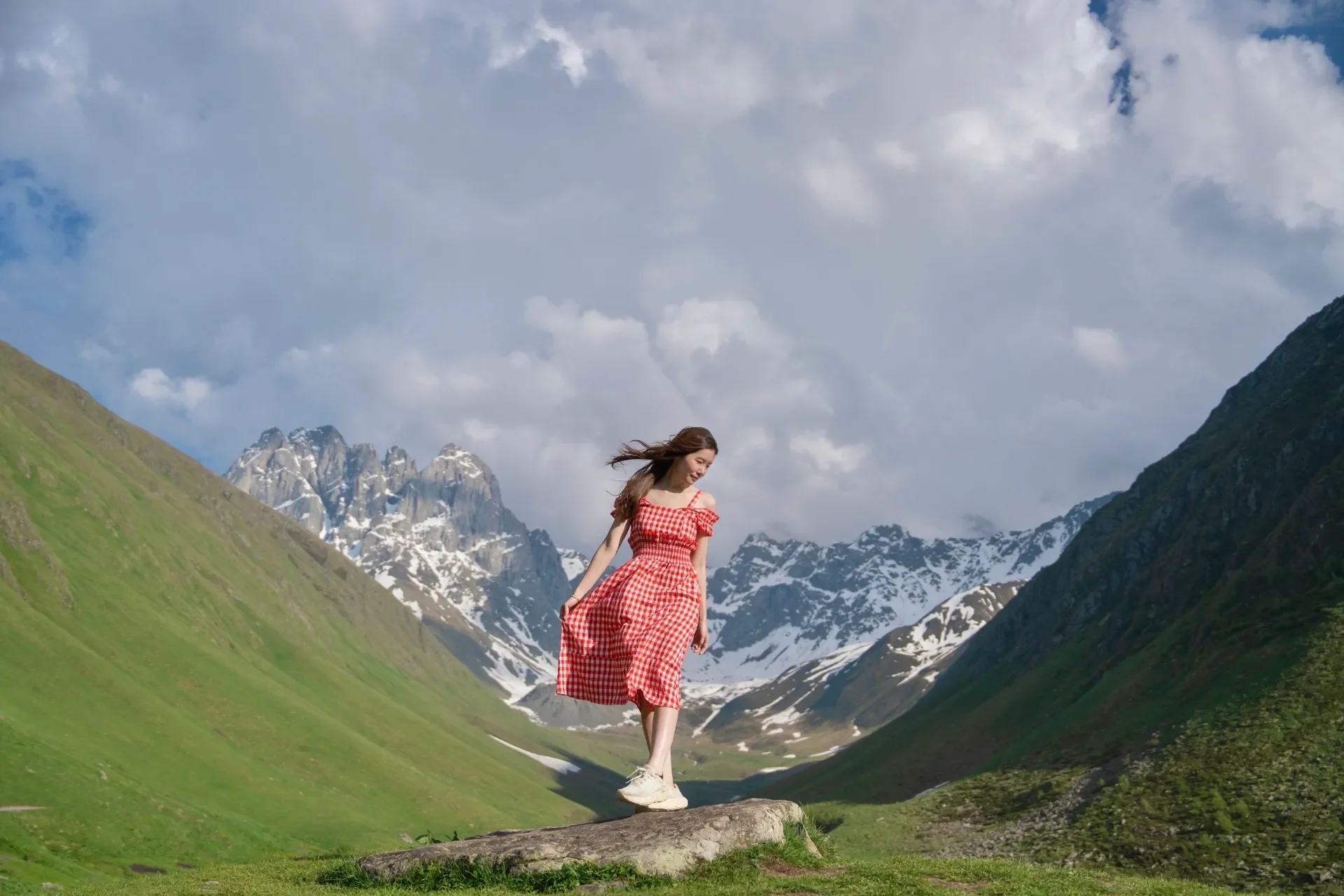 A woman in a red checkered dress standing on a large rock in a lush green valley surrounded by snow-capped mountains.