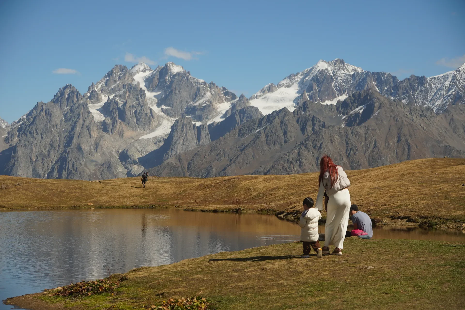 People near a small lake with mountains in the background, including a woman and a child standing by the water, and another person sitting by the lake.