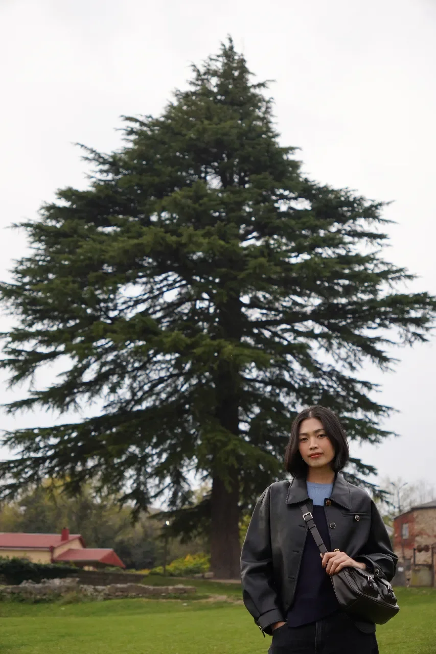 A woman standing in front of a large pine tree, wearing a black leather jacket and carrying a black handbag outdoors on an overcast day