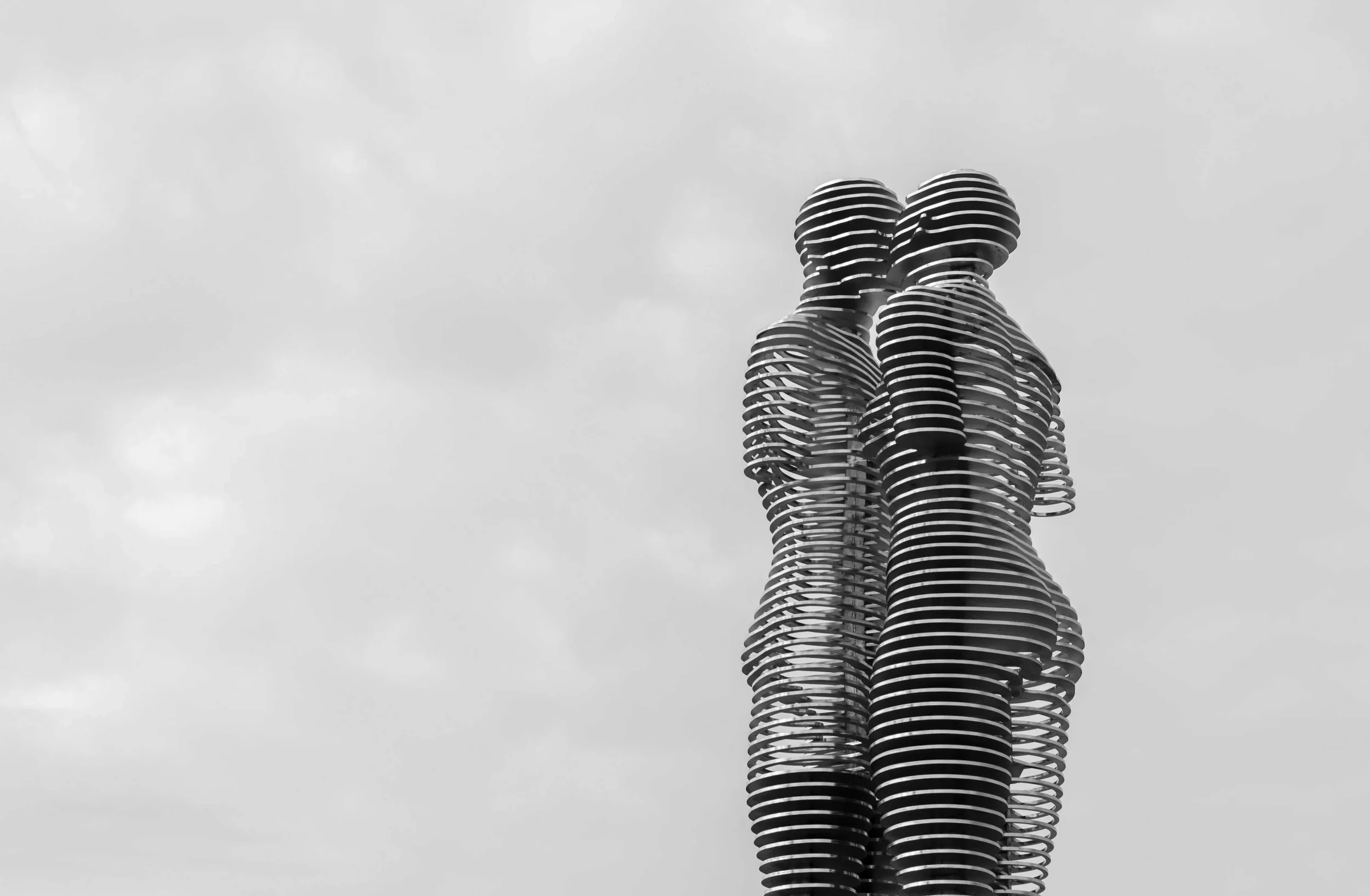 A tall modern sculpture of two people facing each other, made of horizontal layered metal slats, set against a cloudy sky.